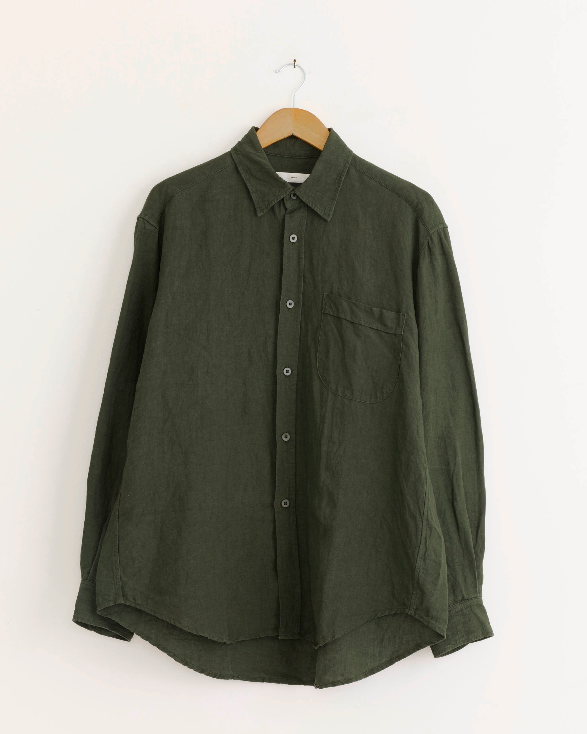 The SMOCK Gio Shirt in Sun Dried Linen Green—a long-sleeved, dark green Japanese linen shirt with a collar and chest pocket, made in Los Angeles—hangs on a wooden hanger against a plain white wall.