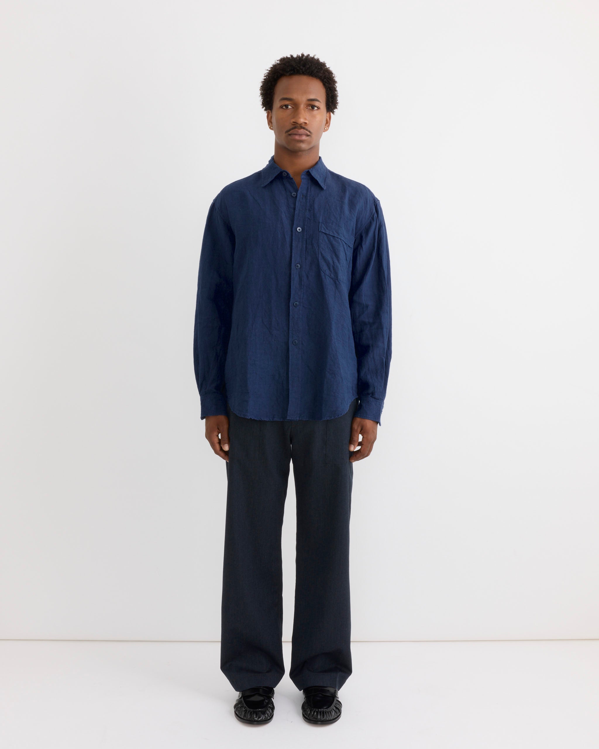 A person stands facing forward against a plain white background, wearing the SMOCK Gio Shirt in Sun Dried Linen Blue— a loose-fitting, long-sleeve Japanese linen shirt—paired with dark wide-leg pants and black shoes.