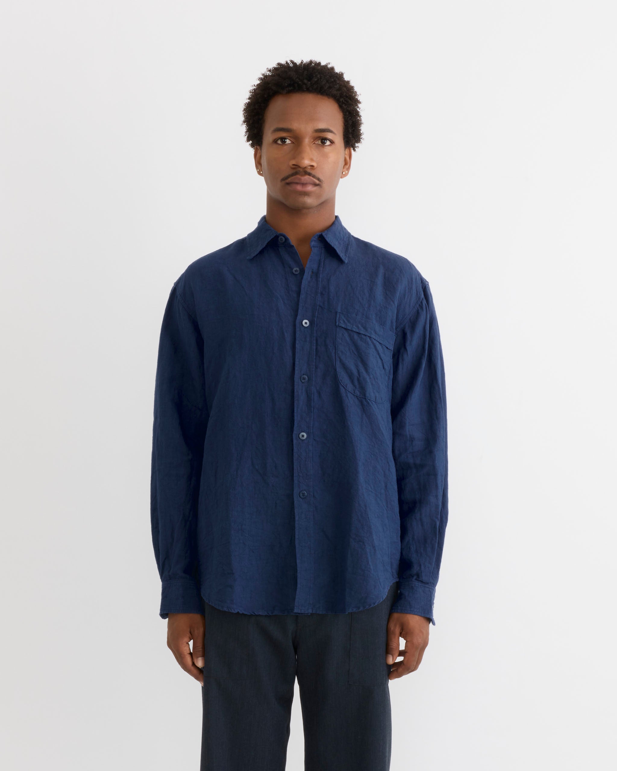 A person stands facing forward against a plain white background, wearing the SMOCK Gio Shirt in Sun Dried Linen Blue and dark pants. They have short curly hair and a neutral expression.