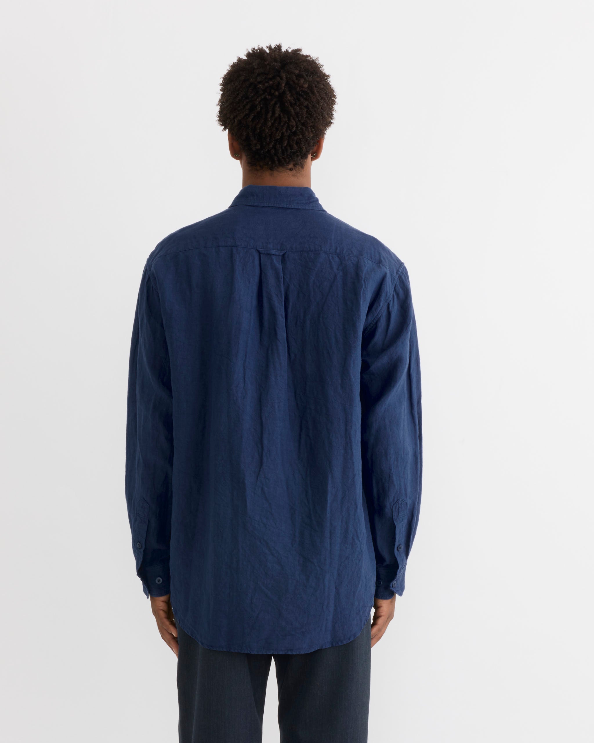 A person stands facing away from the camera, wearing the SMOCK Gio Shirt in Sun Dried Linen Blue—a loose-fitting, long-sleeve linen shirt—paired with dark pants against a plain white background.