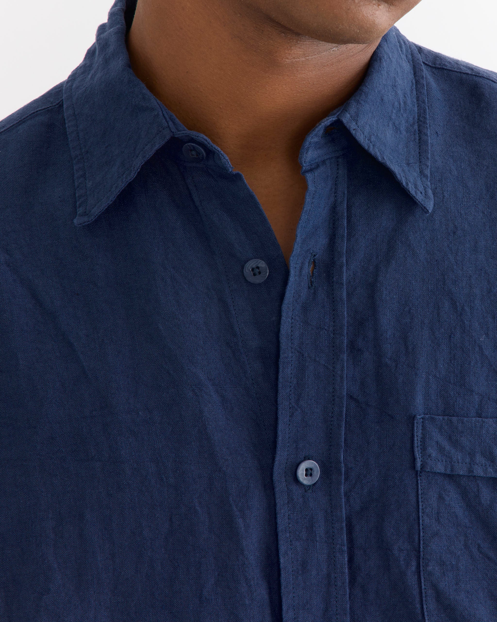 A person wearing the SMOCK Gio Shirt in Sun Dried Linen Blue, featuring a collar and chest pocket, is shown from the shoulders to upper chest with the top button undone.
