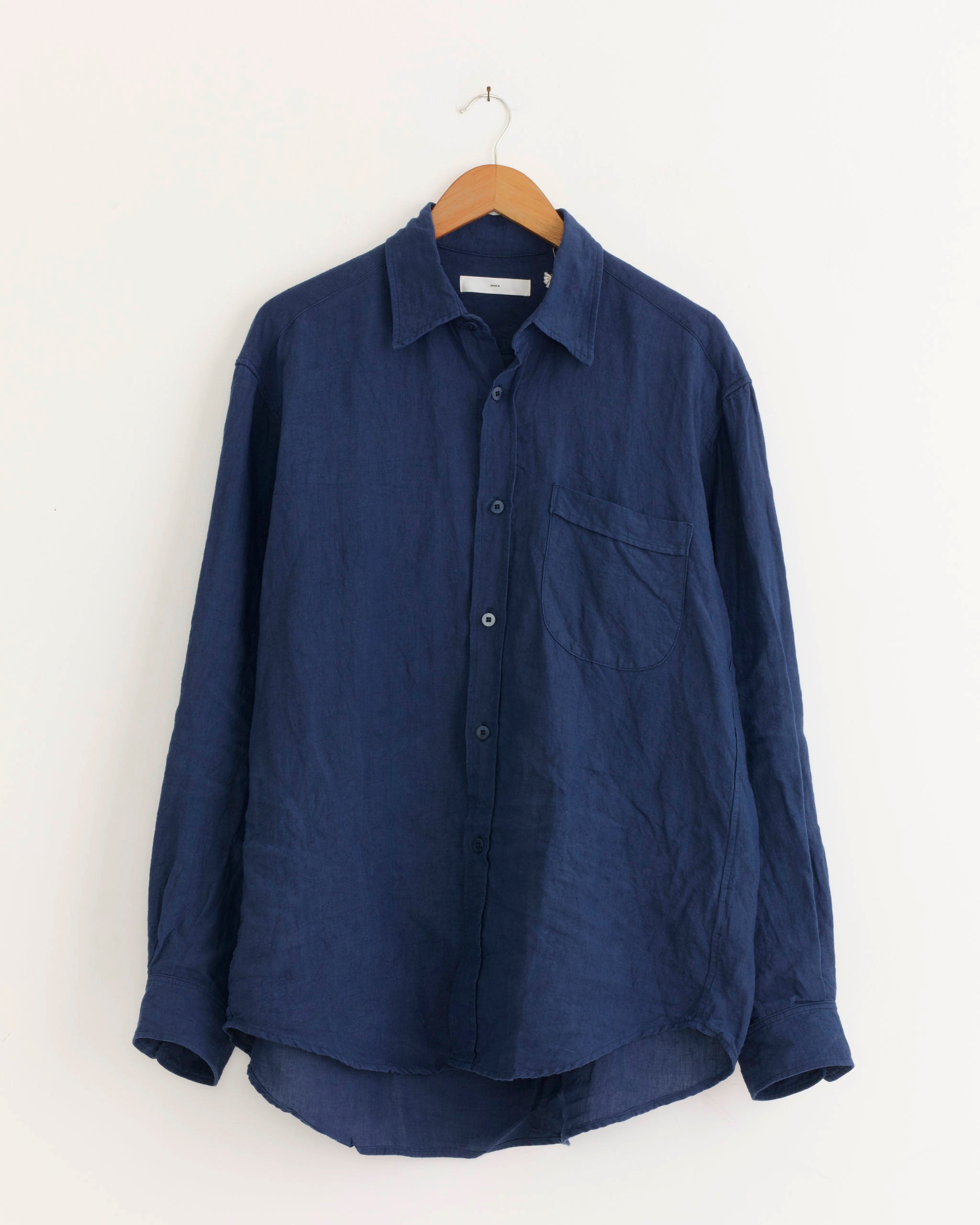 The Gio Shirt in Sun Dried Linen Blue by SMOCK, a long-sleeved, dark blue Japanese linen shirt with a single chest pocket, is hanging on a wooden hanger against a plain white wall.