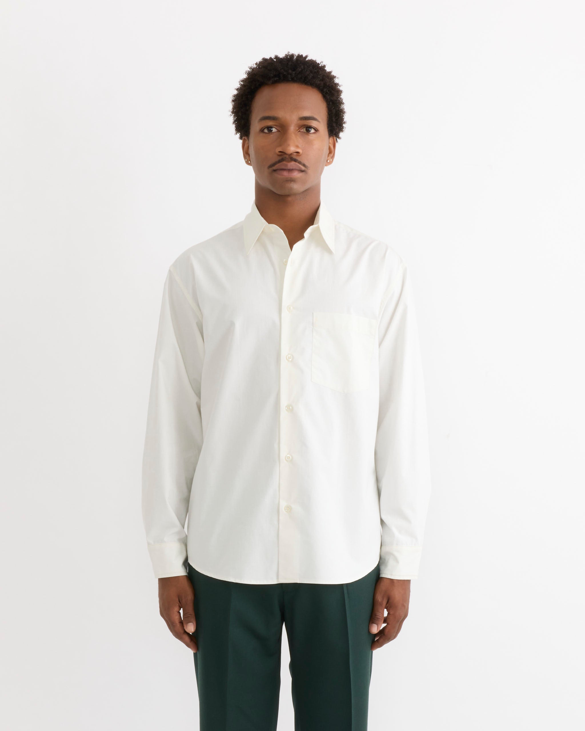 A person stands facing forward against a plain white background, wearing the SMOCK Routine Shirt in Cotton Silk Off White and dark green pants.