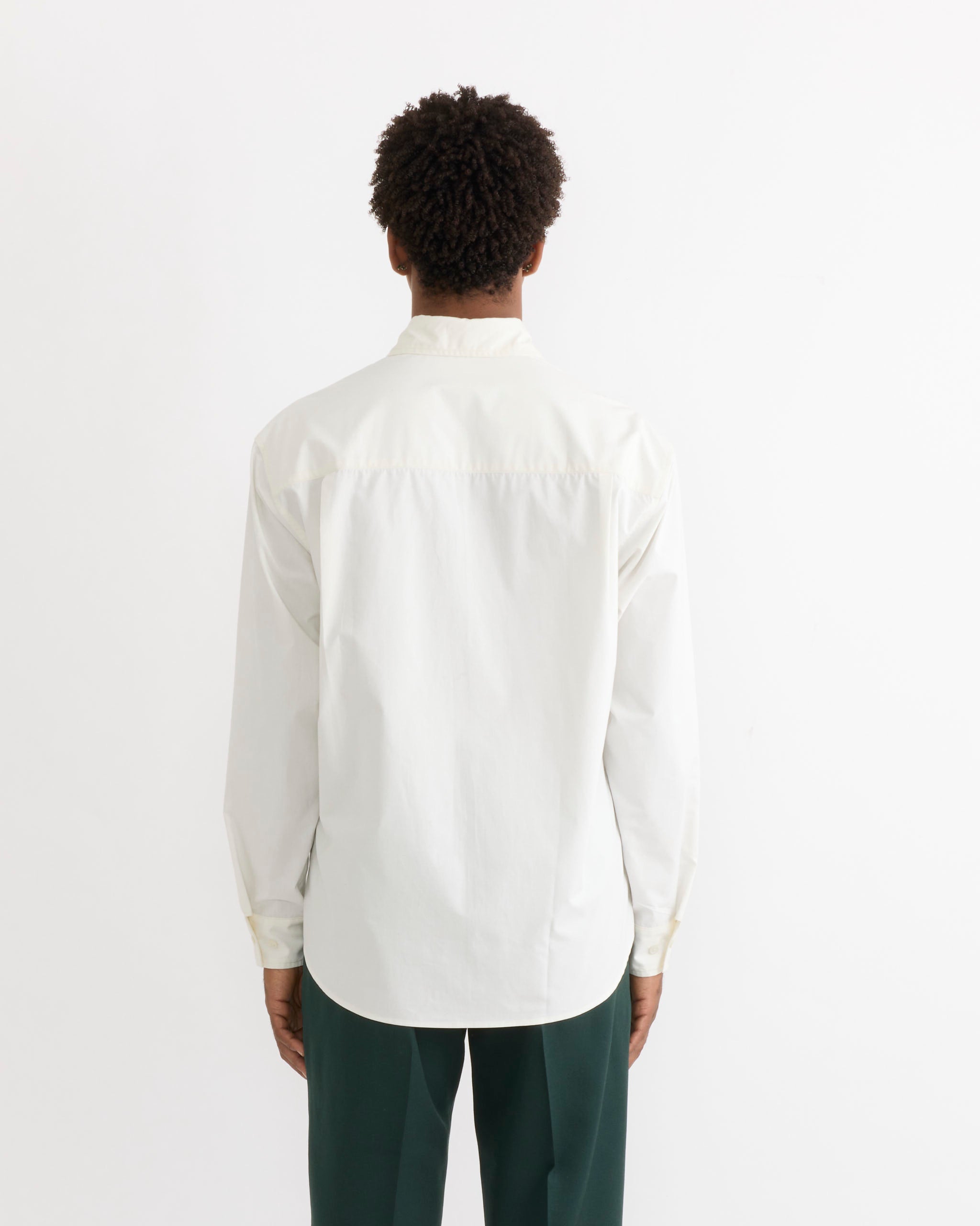 A person stands with their back to the camera, wearing the SMOCK Routine Shirt in Cotton Silk Off White and dark green pants, set against a plain white background.