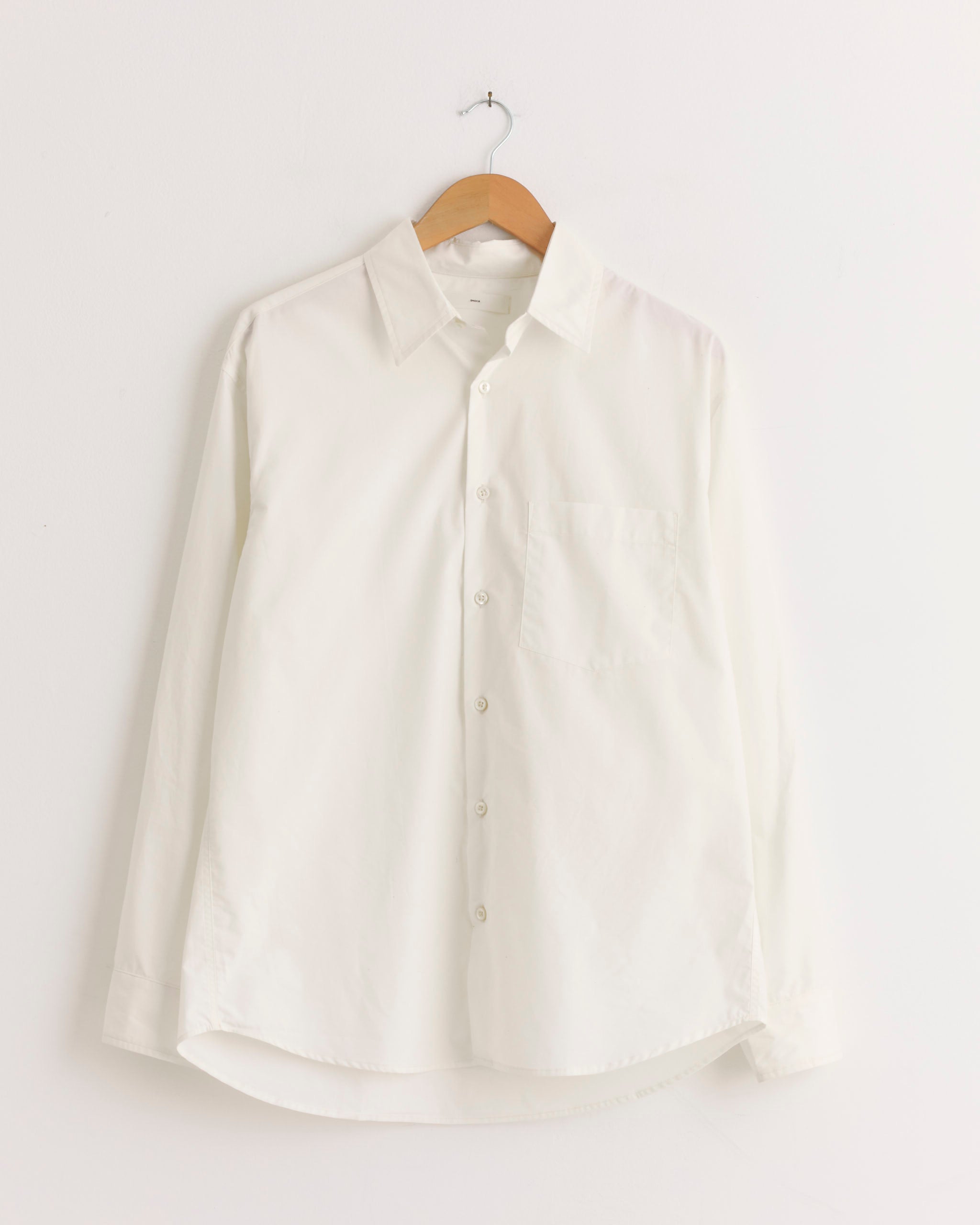 The Routine Shirt in Cotton Silk Off White by SMOCK hangs on a wooden hanger against a white wall.