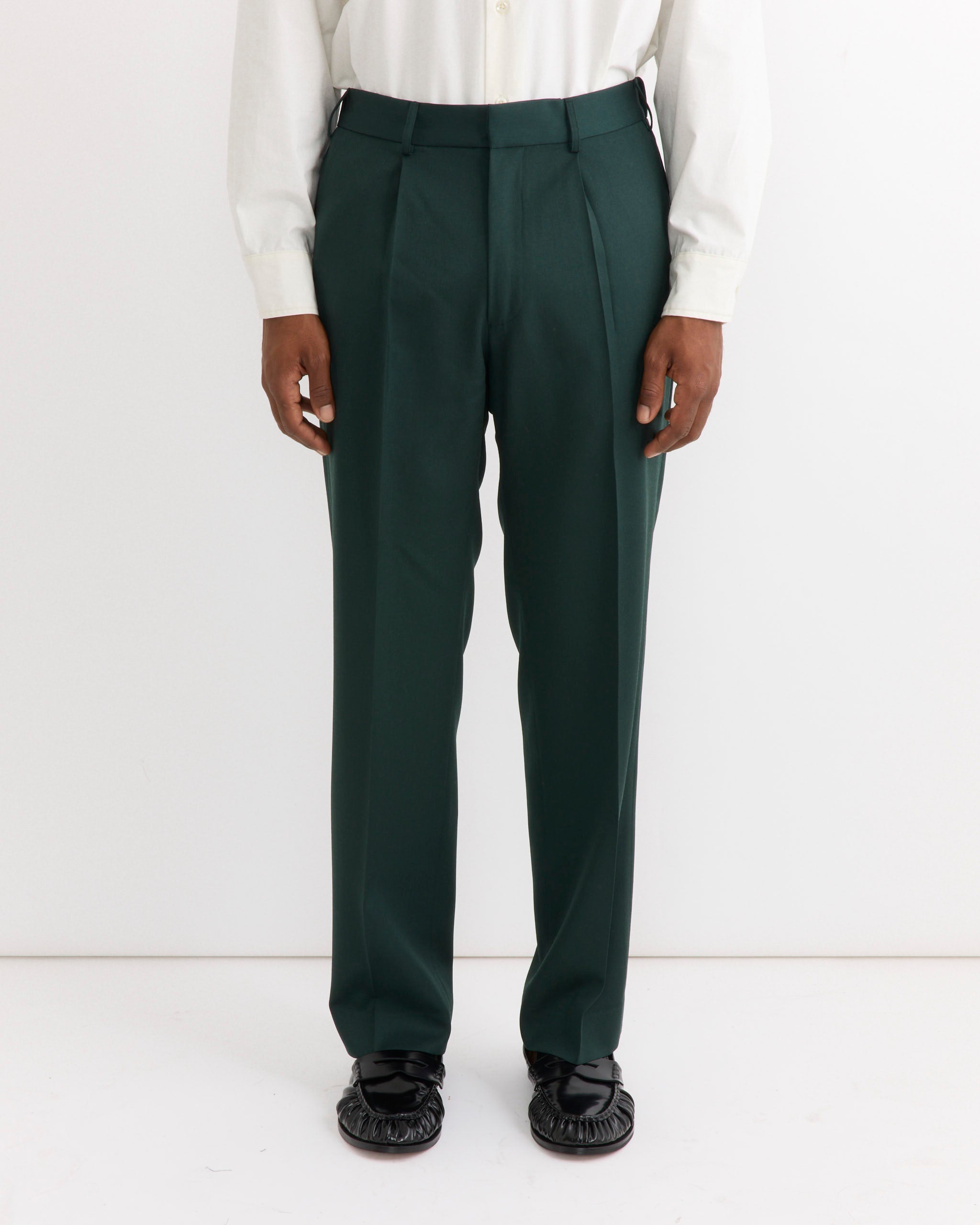 A person wears the SMOCK West Coast Pant in Wool Gabardine Dark Green, paired with a crisp white long-sleeve shirt and black shoes, standing against a plain white background.