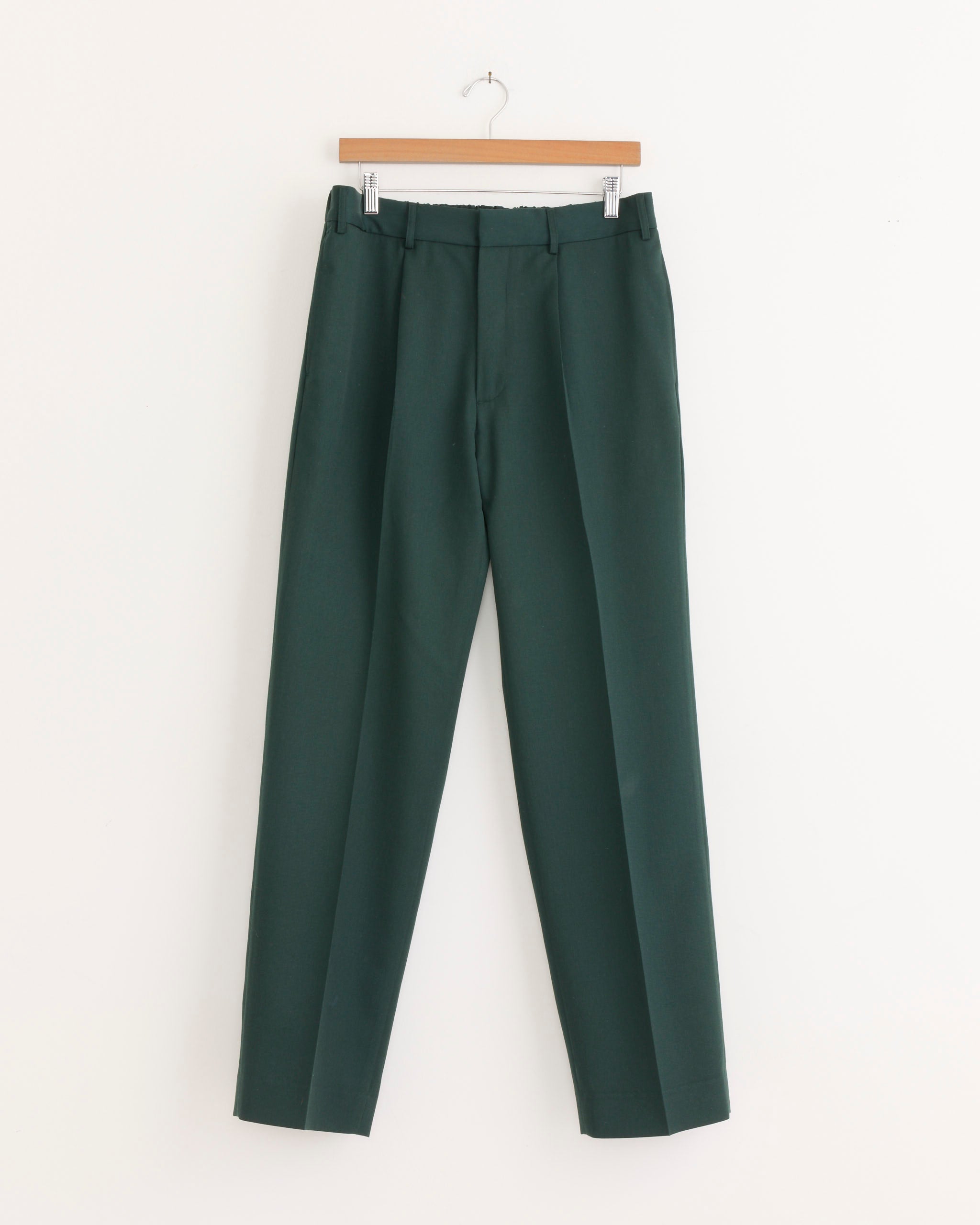 The SMOCK West Coast Pant in Wool Gabardine Dark Green hangs from a wooden hanger against a plain white background. Tailored with belt loops, these trousers feature a straight-leg silhouette and a sharp front crease, crafted from Japanese Wool Gabardine.