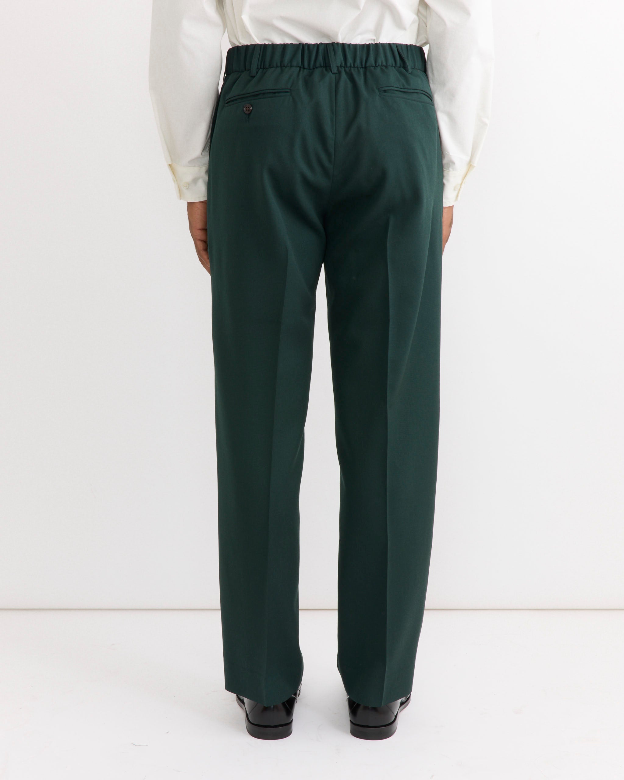 A person is shown from the back wearing the SMOCK West Coast Pant in Wool Gabardine Dark Green, styled with a white shirt. Crafted from Japanese wool gabardine, these trousers feature an elastic waistband and back pockets. The individual stands against a plain white background.