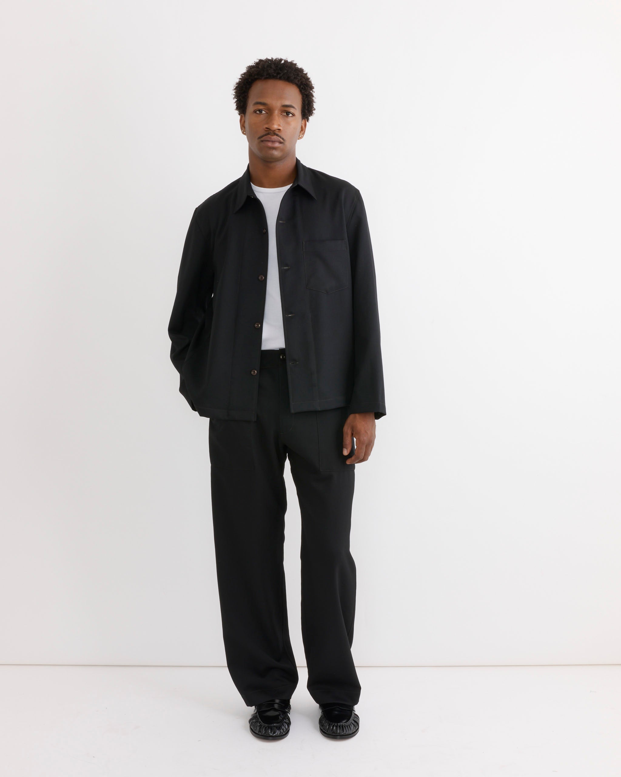 A man stands against a white wall wearing a black jacket, white T-shirt, SMOCK Toyon Pant in Wool Gabardine Black, and black shoes, with one hand in his pocket.