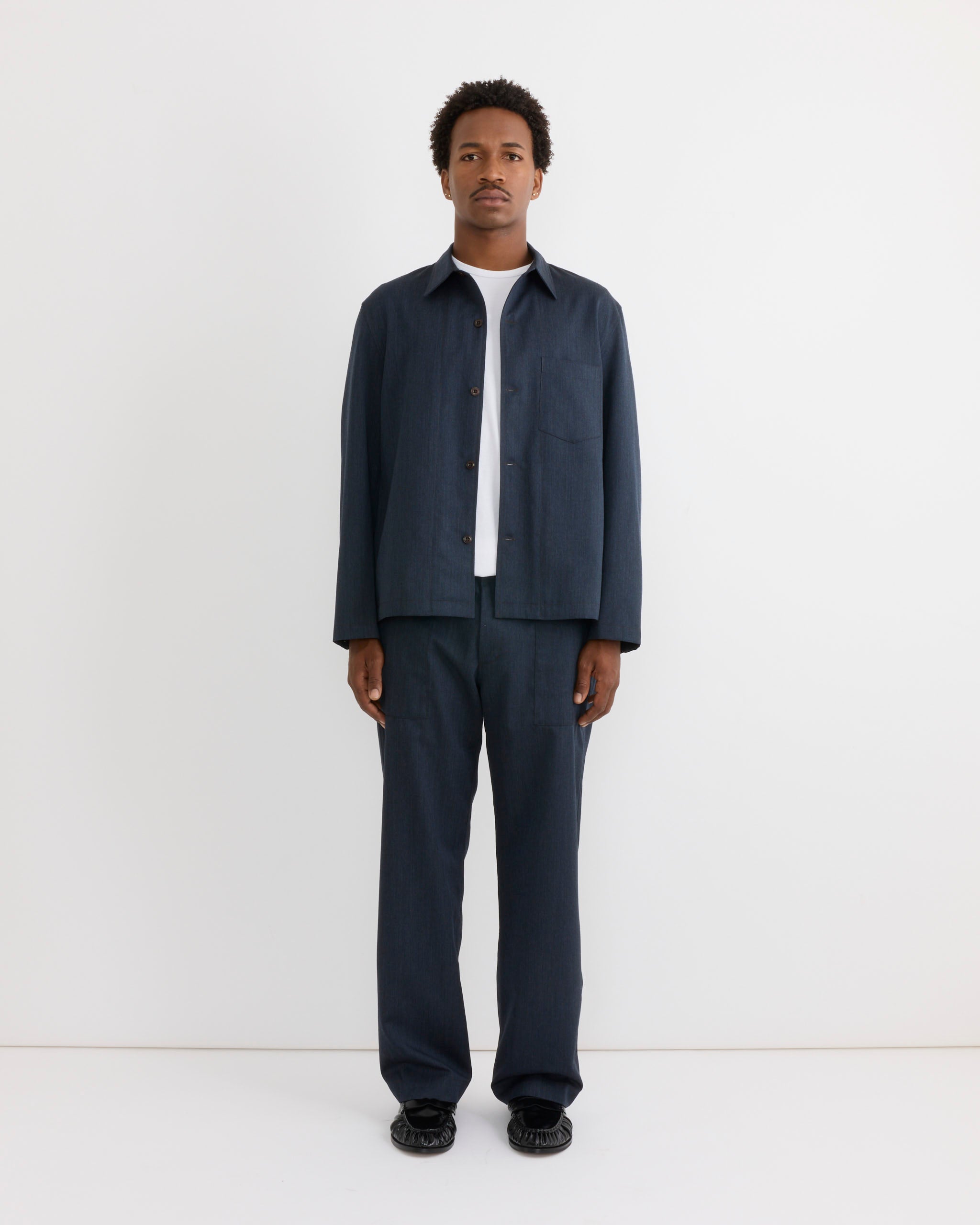 A person stands against a plain white wall, facing forward with arms at their sides, wearing a navy jacket over a white shirt, SMOCK’s Toyon Pant in Wool Gabardine Heather Navy in a relaxed straight-leg style, and black shoes.