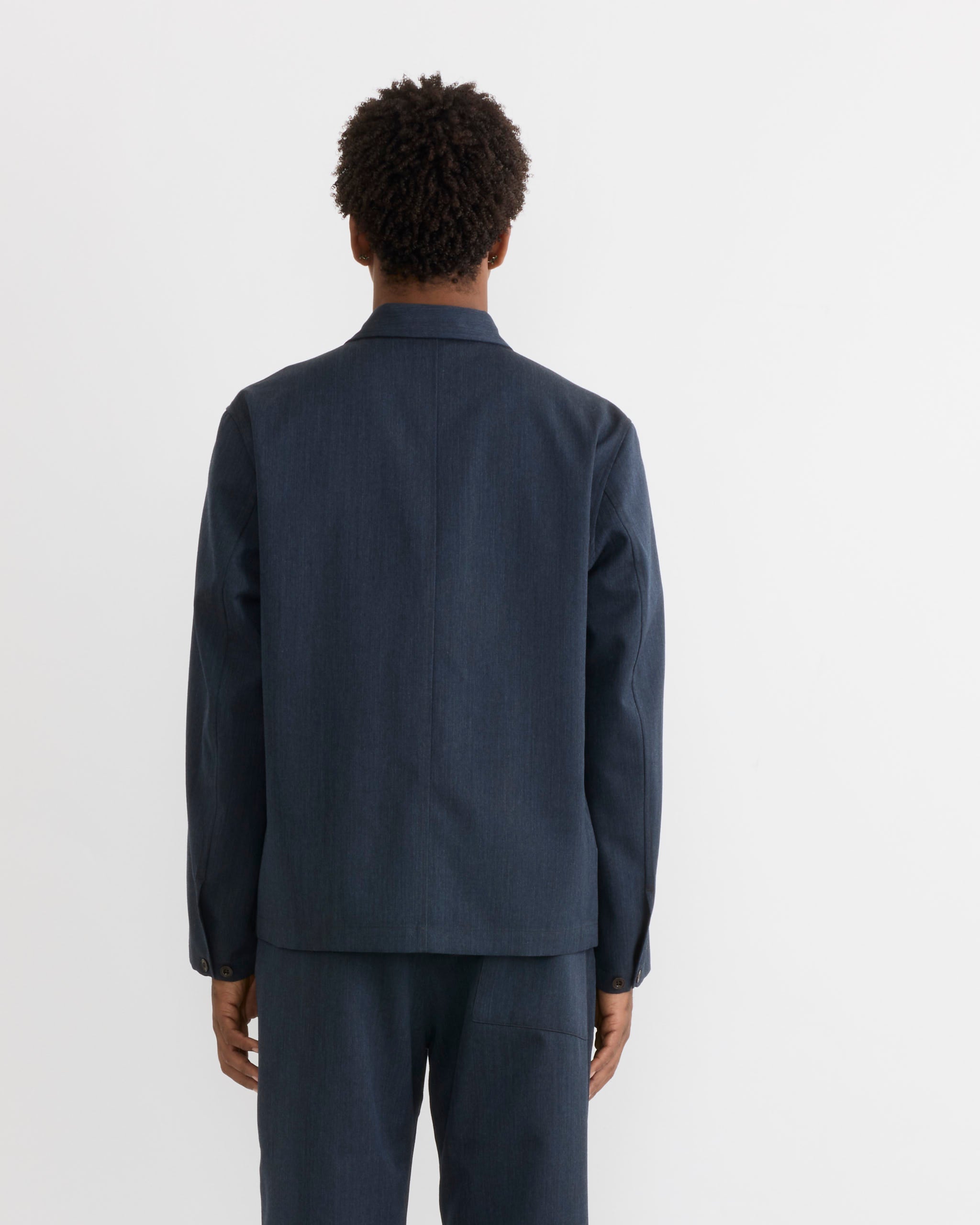 A person with short curly hair, shown from the back against a plain white background, wears a dark blue suit styled formally and tailored for a precise fit. Completing the look is SMOCK’s Notte Shirt in Wool Gabardine Heather Navy, which adds refined texture and elegance to the ensemble. Their arms remain relaxed at their sides, emphasizing the understated sophistication of this well-curated outfit.