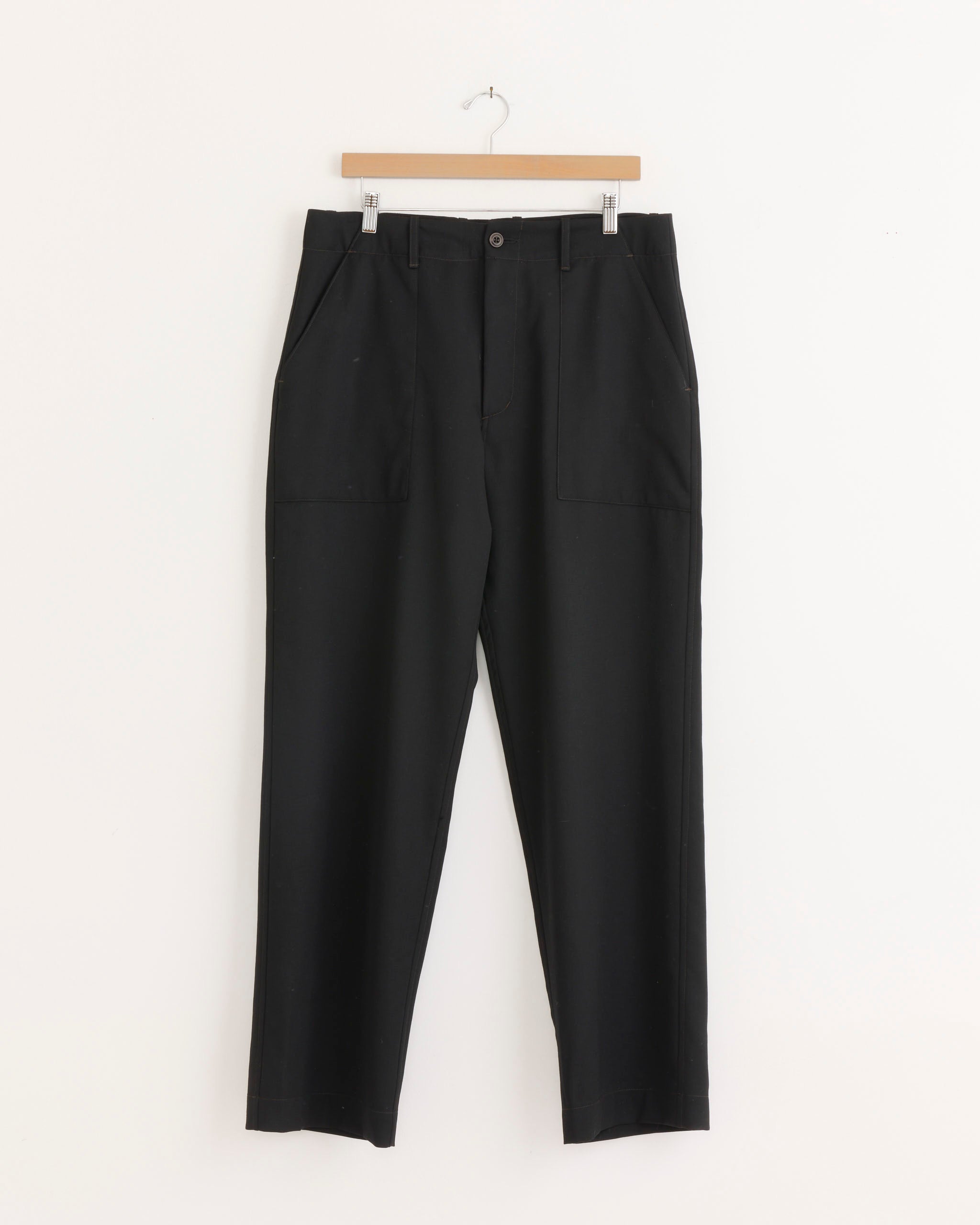 The Toyon Pant in Wool Gabardine Black by SMOCK is a relaxed straight-leg silhouette, crafted from Japanese wool and displayed on a wooden hanger against a white background. These trousers feature front patch pockets and a button closure at the waistband.