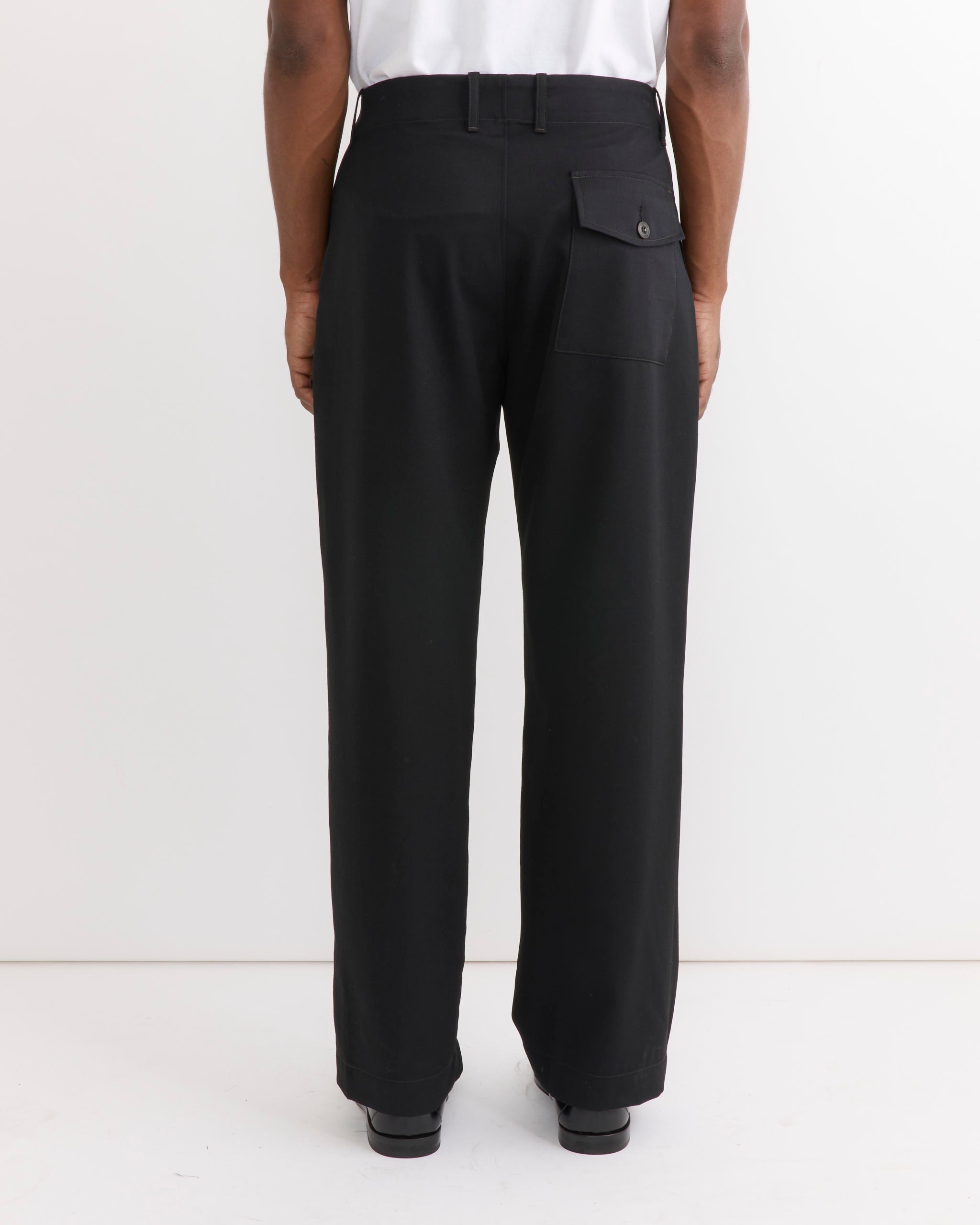 A person stands facing a white wall, wearing SMOCK’s Toyon Pant in Wool Gabardine Black—relaxed straight-leg trousers featuring a back flap pocket—and black shoes. The upper body is only partially visible, but the tailored silhouette of the pants enhances their overall look.