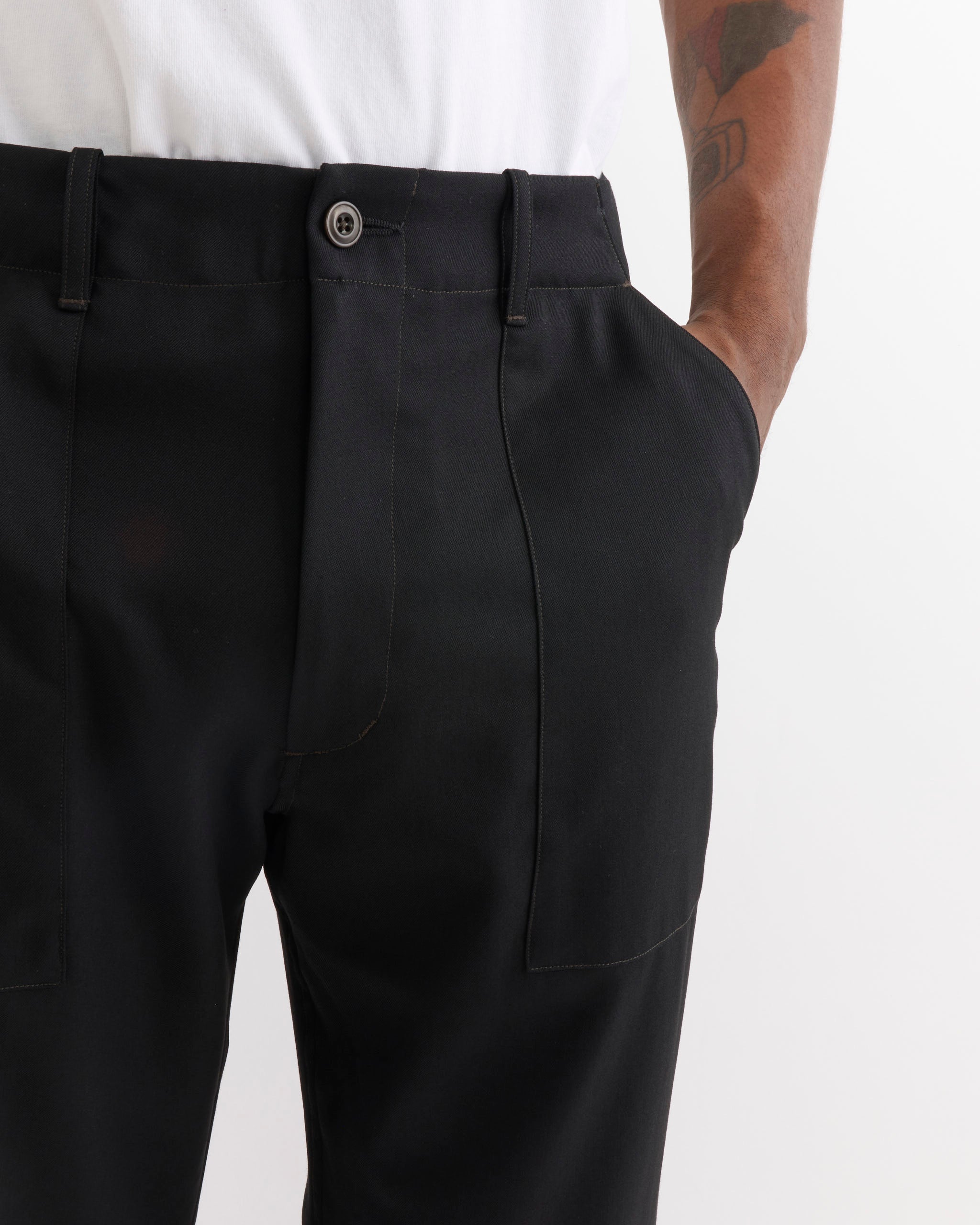A person wearing the SMOCK Toyon Pant in Wool Gabardine Black and a white shirt stands with one hand in their pocket, revealing part of a tattooed arm.