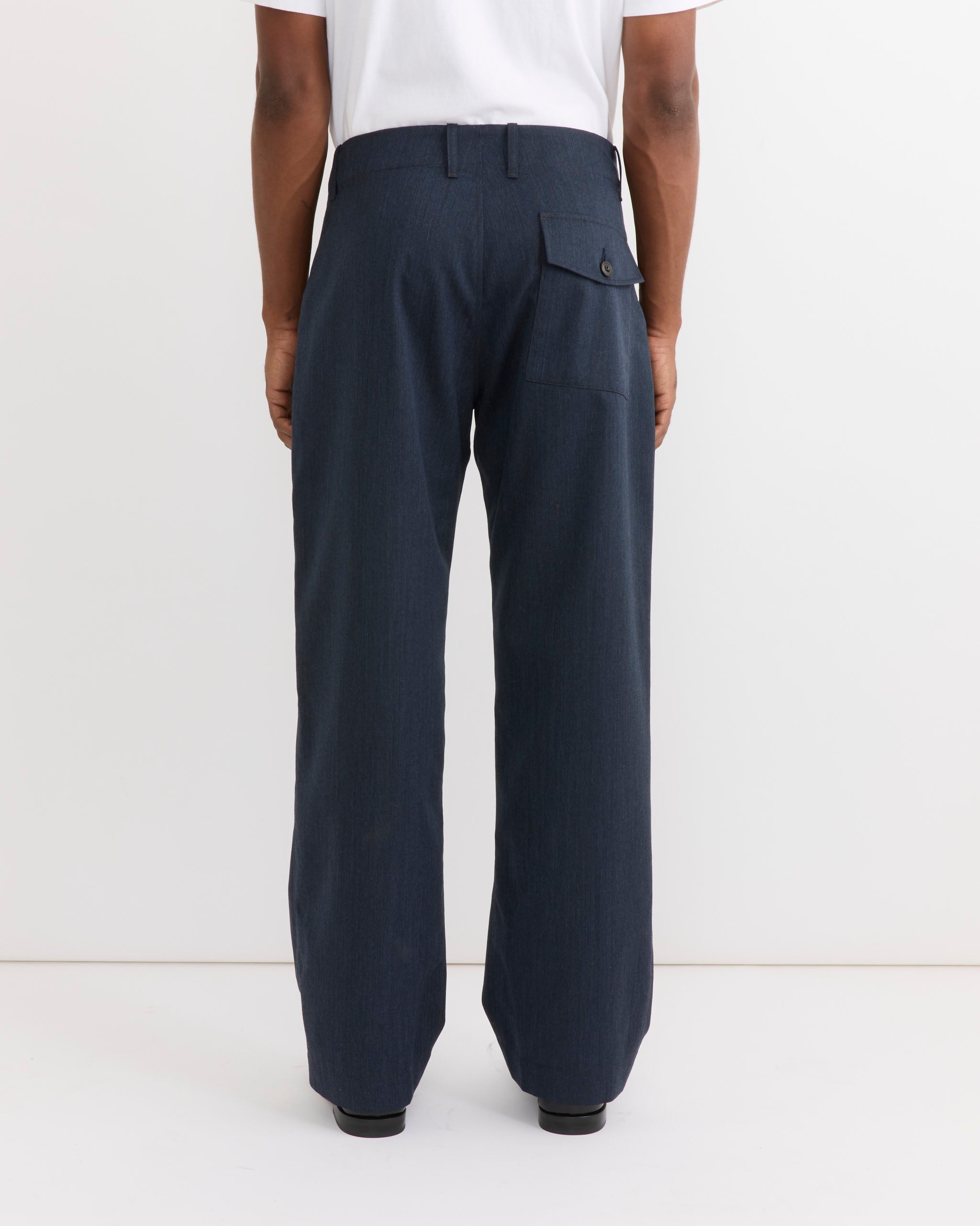 A person stands facing away, wearing the SMOCK Toyon Pant in Wool Gabardine Heather Navy with a side flap pocket and a white shirt. The background is plain white.