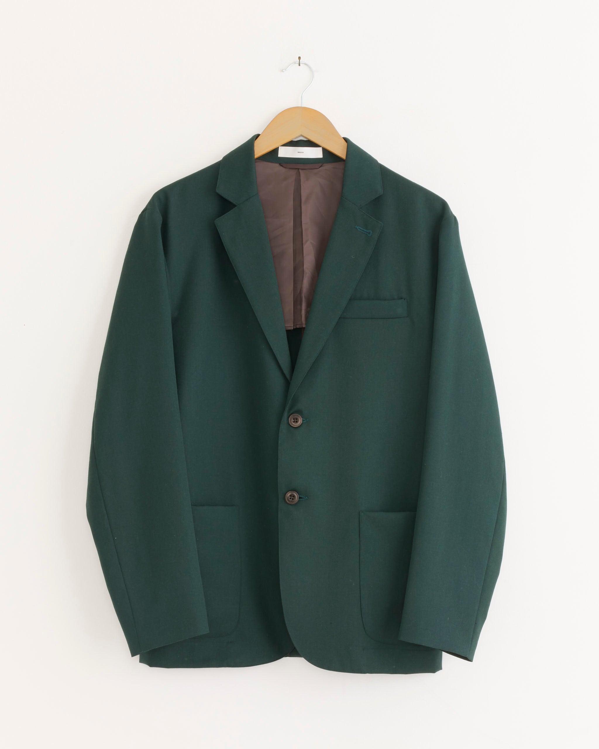 A Blazer in Wool Gabardine Dark Green by SMOCK, featuring a tailored Los Angeles-inspired design with two buttons and two front pockets, is displayed on a wooden hanger against a plain white background.