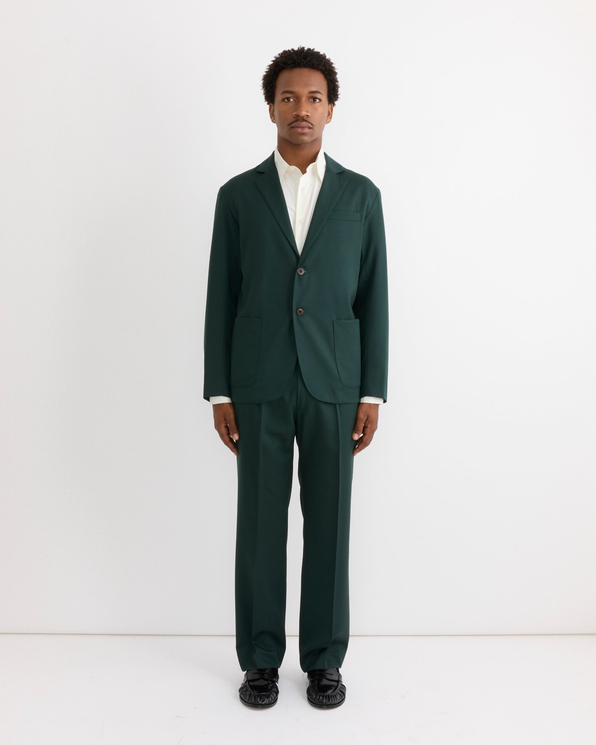 A person stands against a white background wearing the SMOCK Routine Shirt in Cotton Silk Off White, paired with a dark green suit and black shoes, looking straight ahead with a neutral expression.