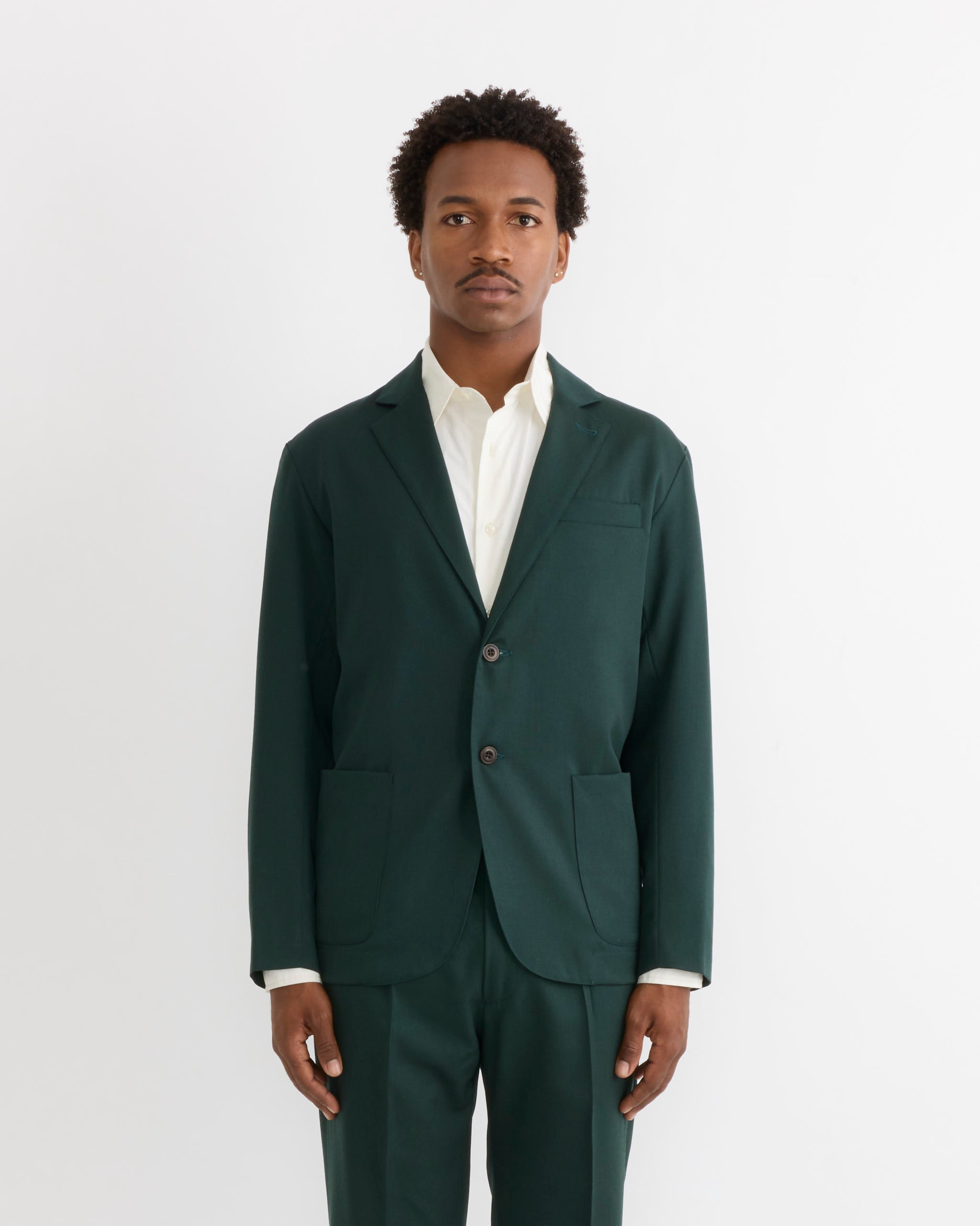 A person stands facing forward, wearing the SMOCK Blazer in Wool Gabardine Dark Green and matching pants over a white dress shirt, against a plain white background.