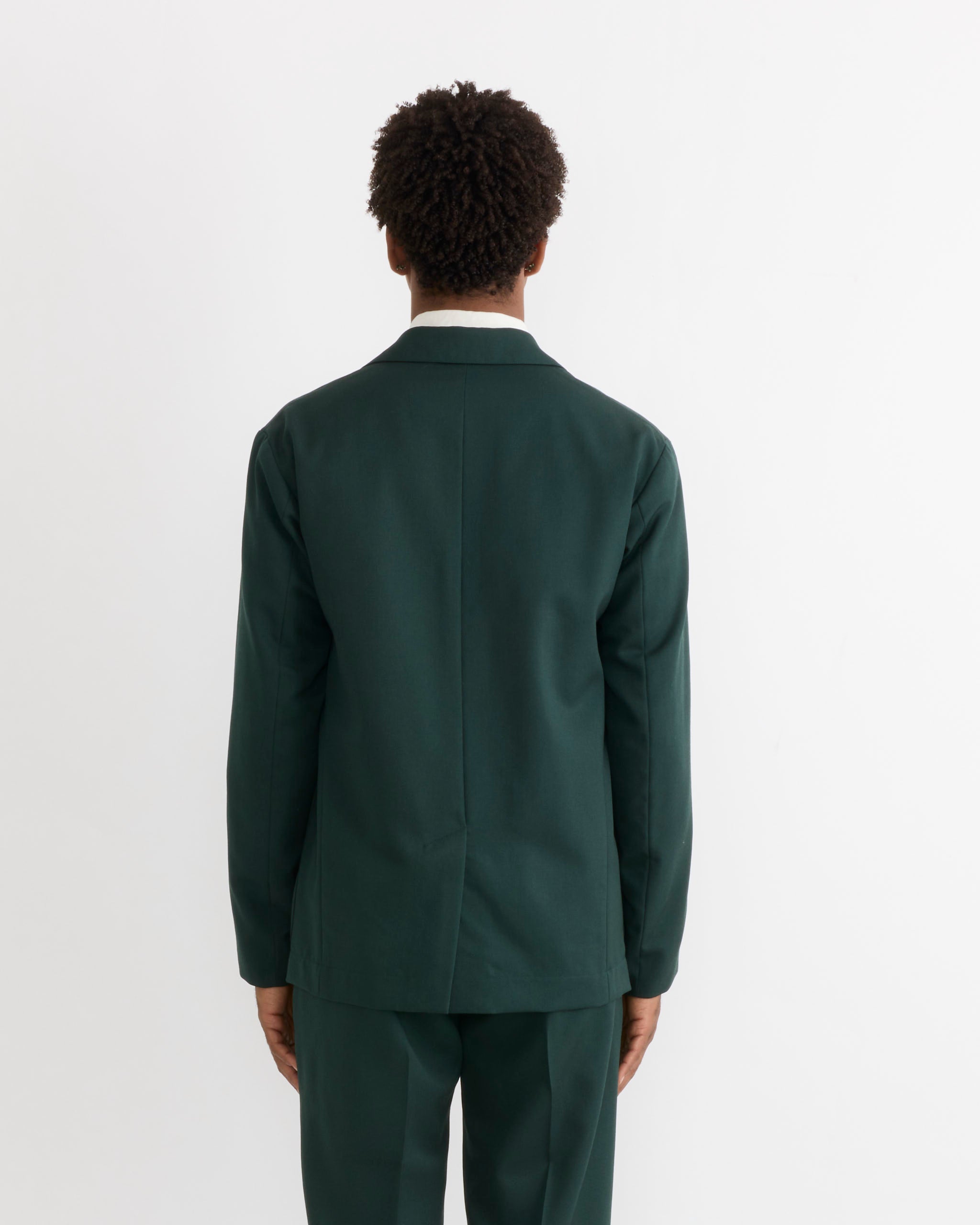 A person with short curly hair, seen from behind, wears the SMOCK Blazer in Wool Gabardine Dark Green and matching pants against a plain white background.