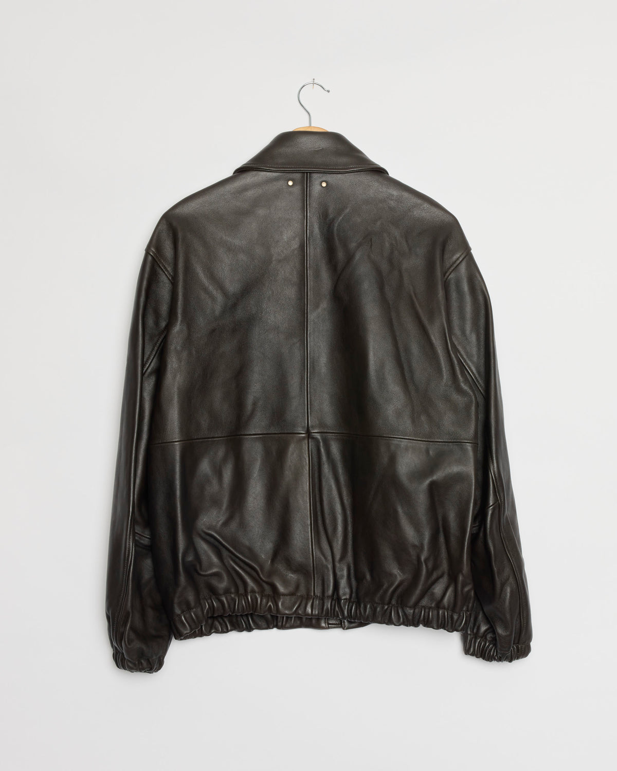 The 1555 Jacket in Vintage Black by ssstein, crafted from supple sheep leather, hangs on a metal hanger and is displayed from the back against a plain white wall.