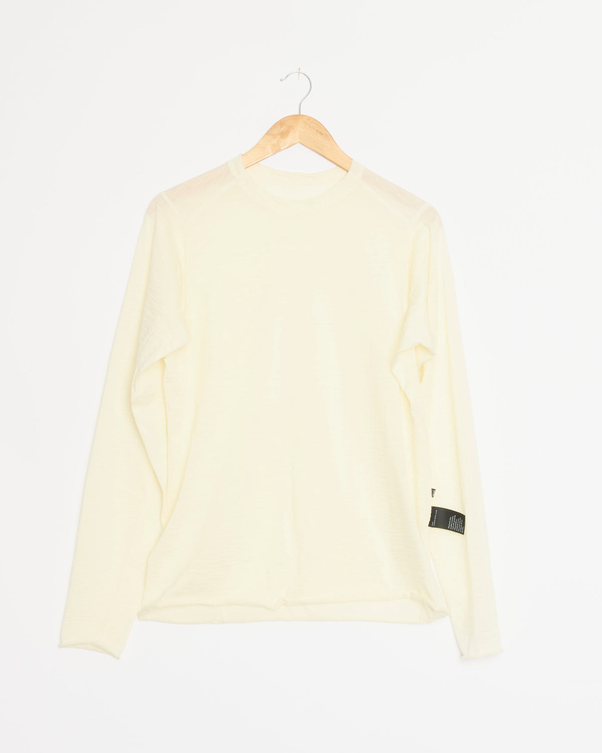 A long-sleeve shirt in the shade “Natural” from Shade, crafted from Italian-milled merino wool, hangs on a wooden hanger against a white background. A small black tag accents one sleeve.