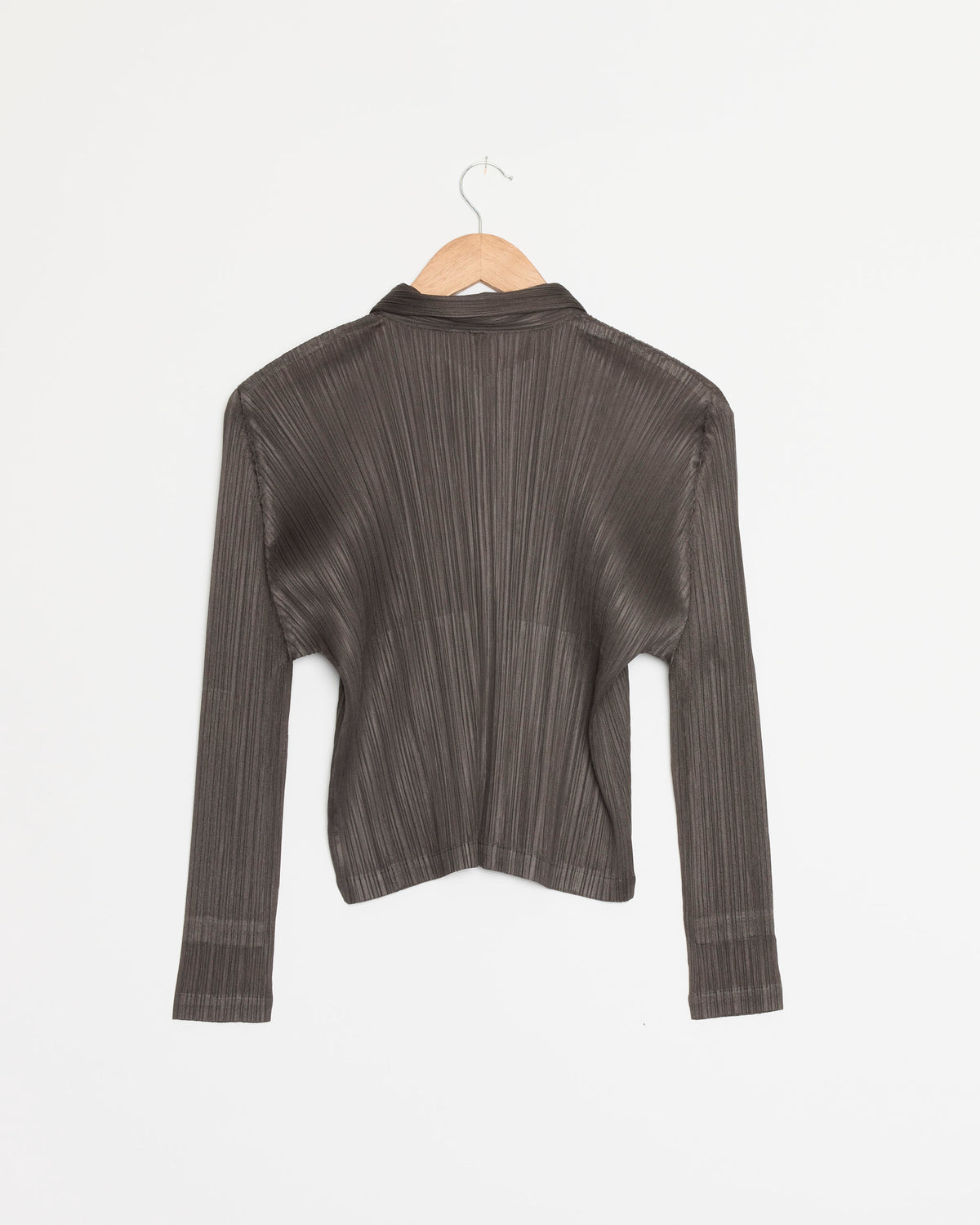 The Monthly Colors August Top in Charcoal by Pleats Please Issey Miyake, a long-sleeved pleated shirt, hangs on a wooden hanger against a plain white background.
