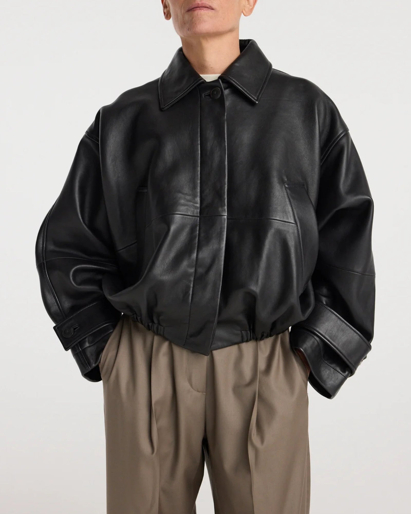 A person wearing the Róhe Leather Blouson Jacket in Black, styled with beige pleated pants, stands against a plain light background. Their face is partially out of frame.