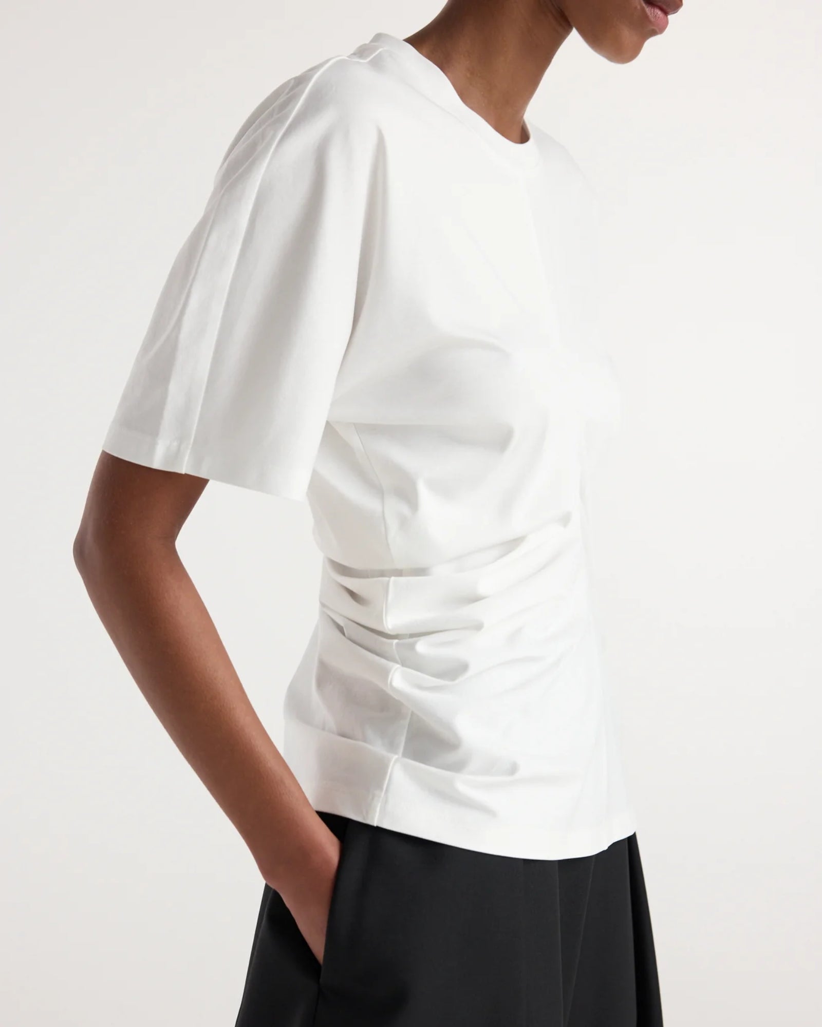 A person is shown from the shoulders to the hips in a side view, standing with hands in pockets against a plain background, wearing the Róhe Draped T-Shirt in Off White and black pants.