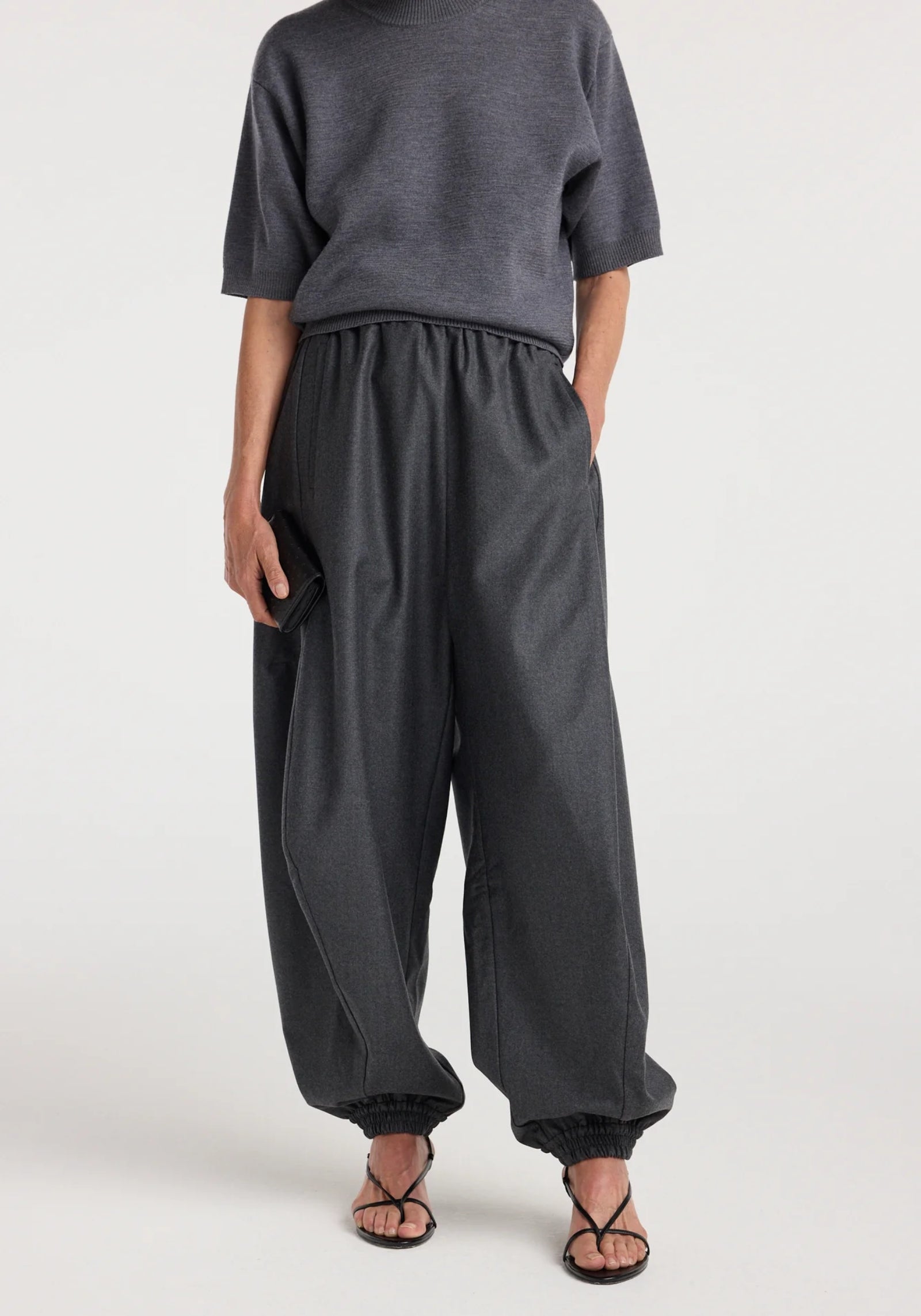 A person wearing the Róhe Balloon Track Pant in Dark Grey Melange—featuring a relaxed fit, elastic waistband, and cuffs—pairs it with a short-sleeve dark gray top and black strappy sandals while standing against a plain background.