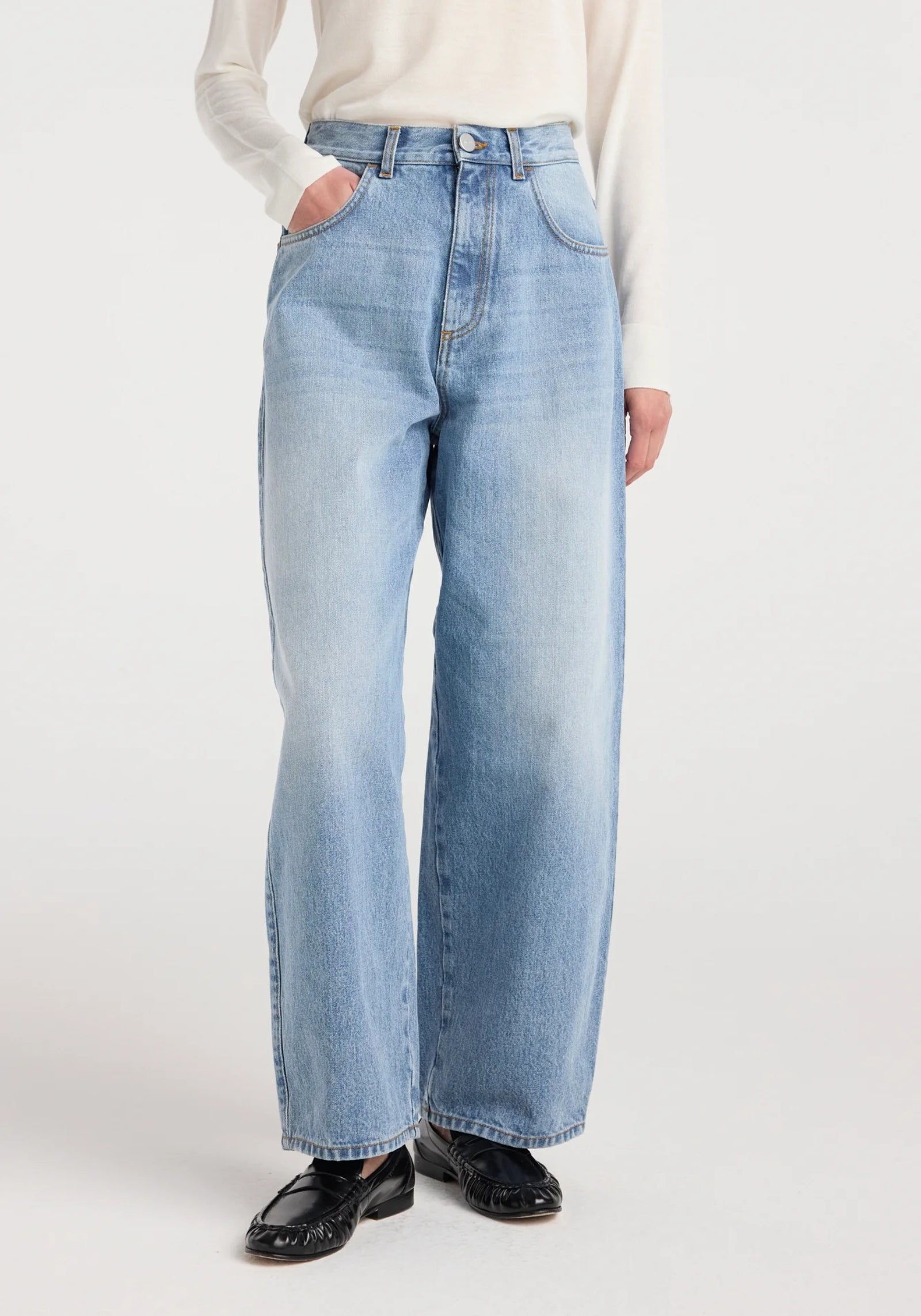 A person wearing the Róhe Berber Barrel Leg Denim Pant in Classic Blue, styled with a tucked-in long-sleeve white shirt and black shoes, stands with one hand in their pocket against a plain white background.