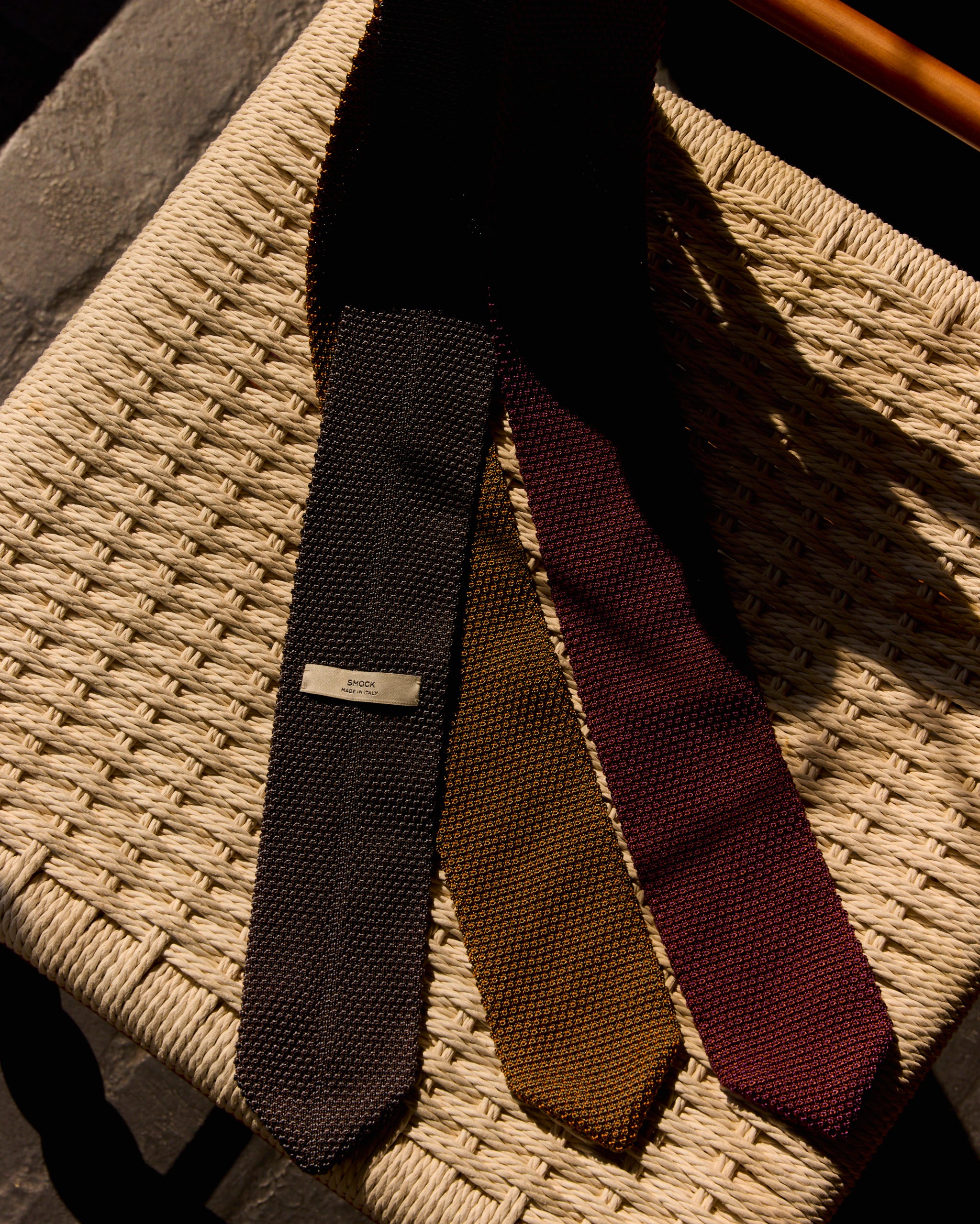 The SMOCK Silk Knit Tie in Olive/Gold—100% silk and made in Italy—rests on a woven straw chair seat, sunlight casting soft shadows across the scene.