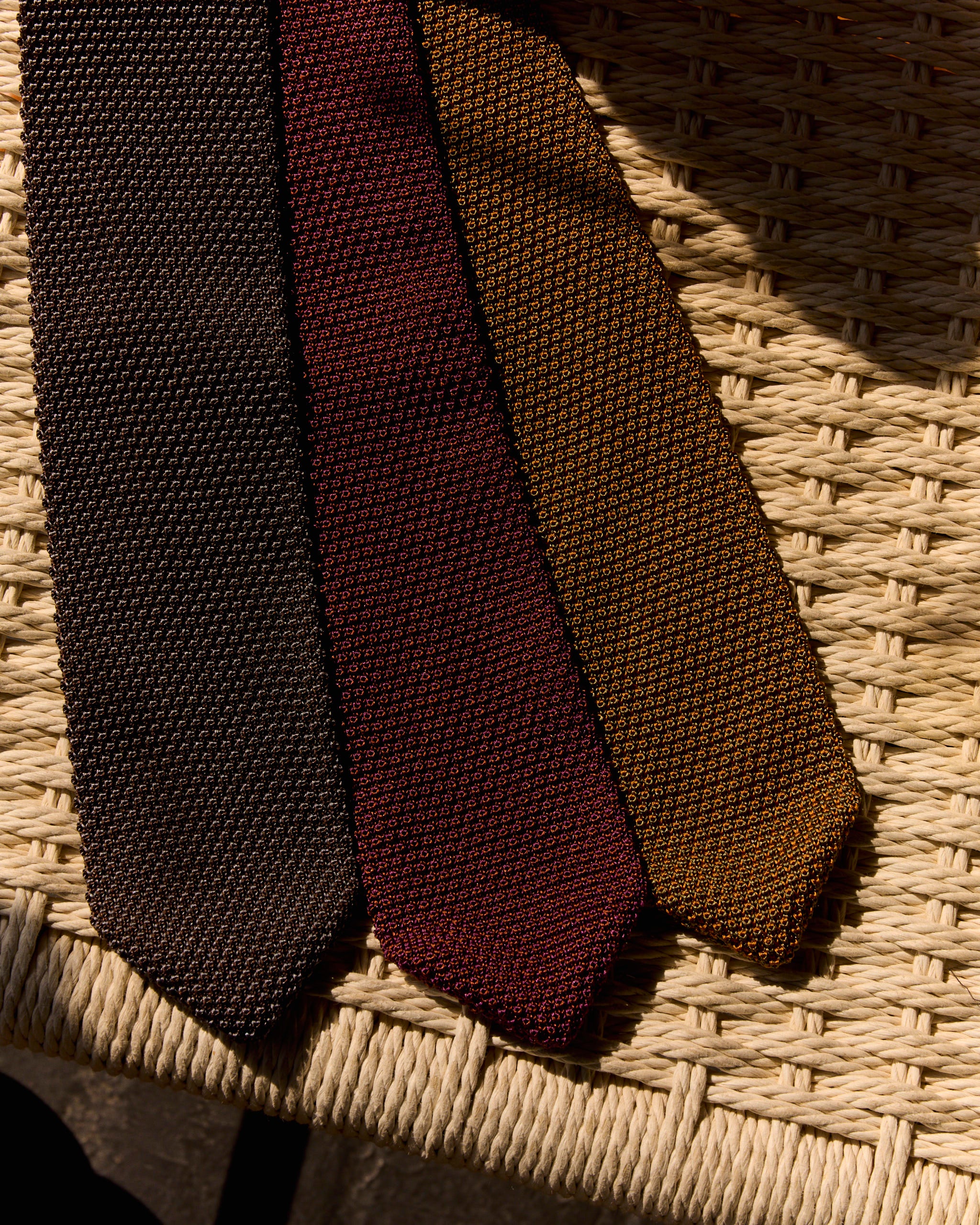 The SMOCK Silk Knit Tie in Eggplant/Cognac, circular knitted and expertly made in Italy, is shown laid on a woven rattan chair under warm lighting.
