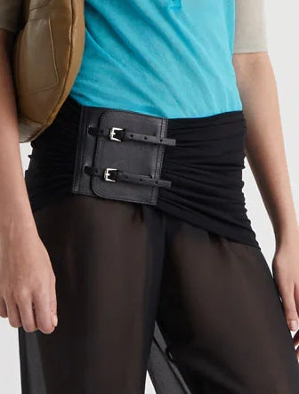 A person wearing a bright blue top, black sheer pants, and the Paloma Wool Adriana II Belt in Multi with double metal buckles at the waist, shown from shoulders to mid-thigh. They carry a tan bag over one shoulder.