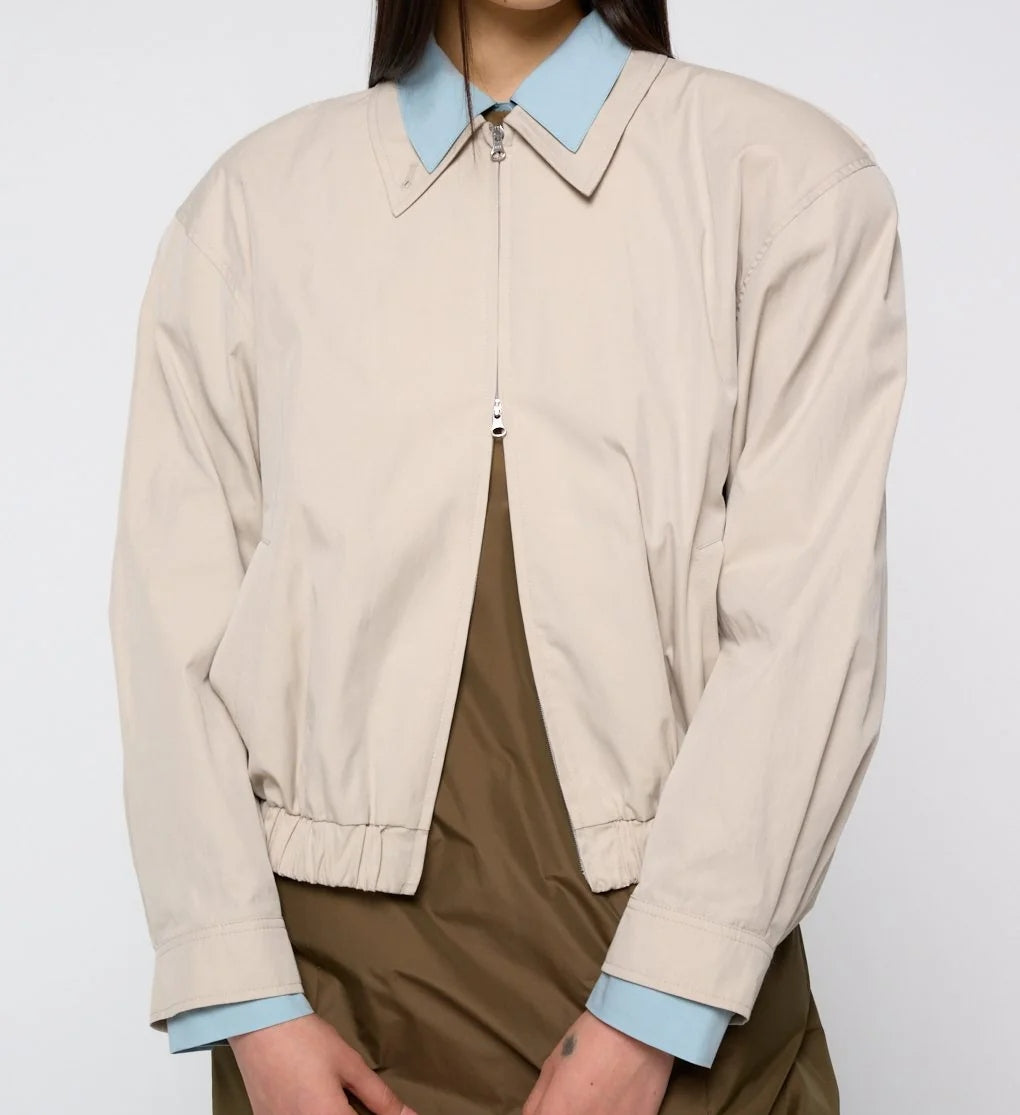 A person wearing the Amomento Cotton Nylon Blouson in Beige over a light blue collared shirt and a brown dress, standing against a plain light background.