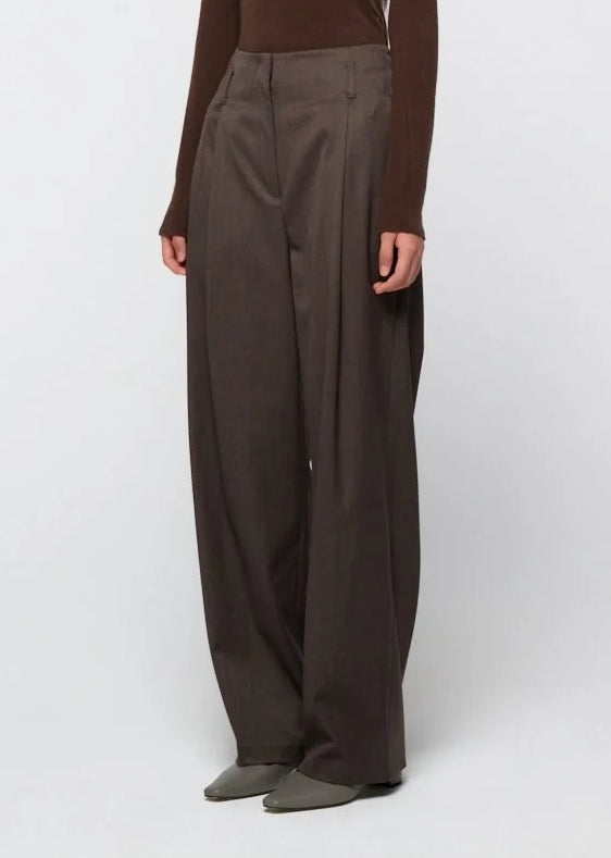 A person is shown standing against a plain light background, wearing Amomento’s Straight Tuck Pant in Brown—a high-waisted, wide-leg wool pant featuring pleats, relaxed silhouette, and subtle tuck detailing—paired with a long-sleeved brown top. The person’s face is not visible.