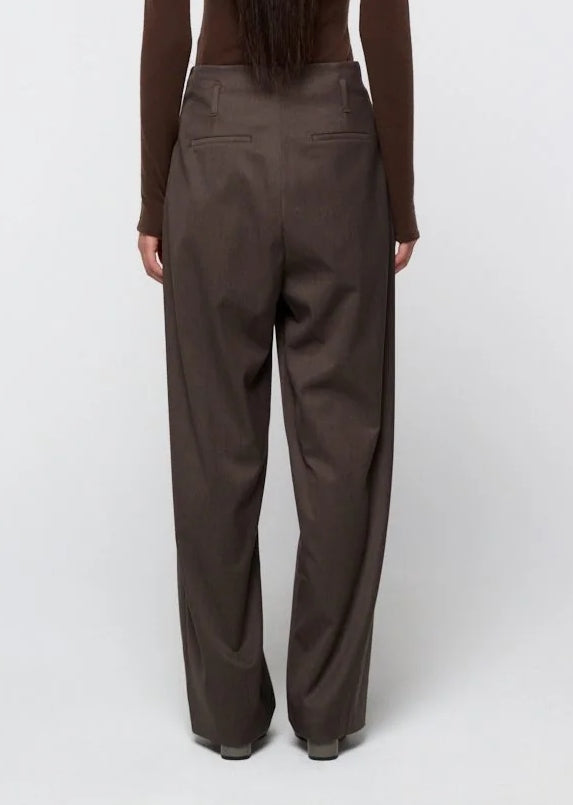 A person stands facing away, dressed in Amomento’s Straight Tuck Pant in Brown—a high-waisted, loose-fitting wool pant with tuck detailing, wide legs, and back welt pockets—paired with a long-sleeve brown top. The background is plain and light.