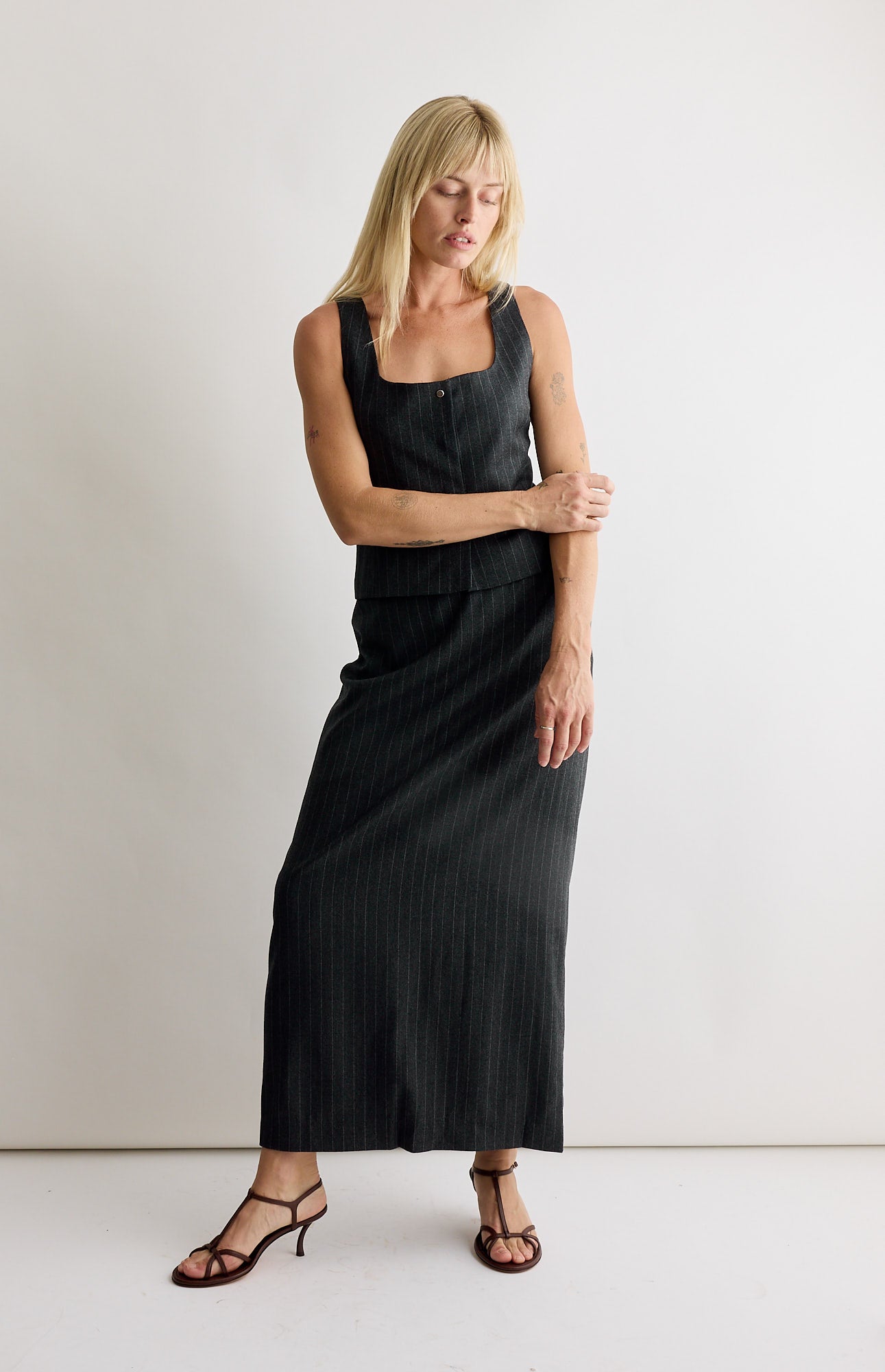 A blonde woman stands against a white background wearing a sleeveless, square-neck black dress with subtle pinstripes inspired by the Amomento Wool Maxi Long Skirt in Stripe and brown strappy sandals.