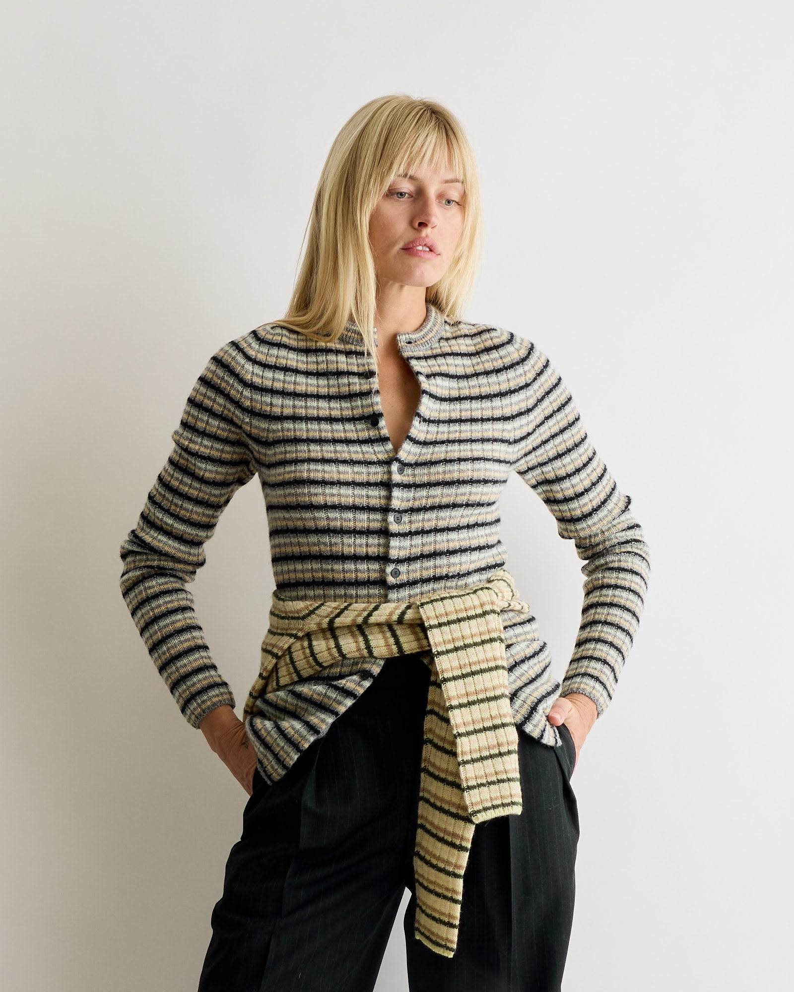 A woman with blonde hair wears the Auralee Wool Cashmere Cardigan in Grey Stripe, a plaid shirt tied at her waist. She stands against a plain white background, hands in pockets, gazing to the side.
