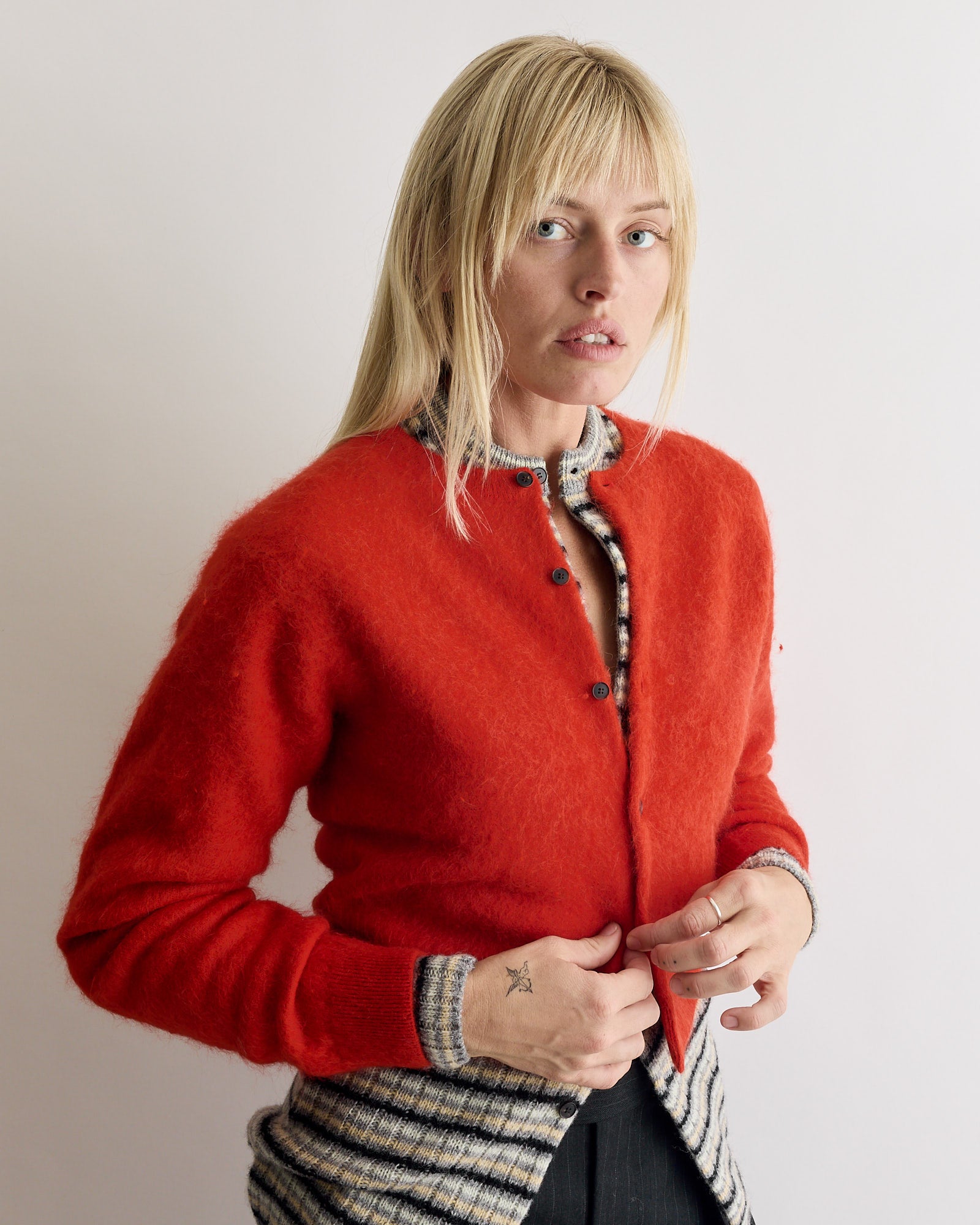 A person with long blonde hair wears the Auralee Baby Alpaca Cardigan in Red over a black-and-white checkered shirt, standing against a plain light background and looking at the camera with a neutral expression.