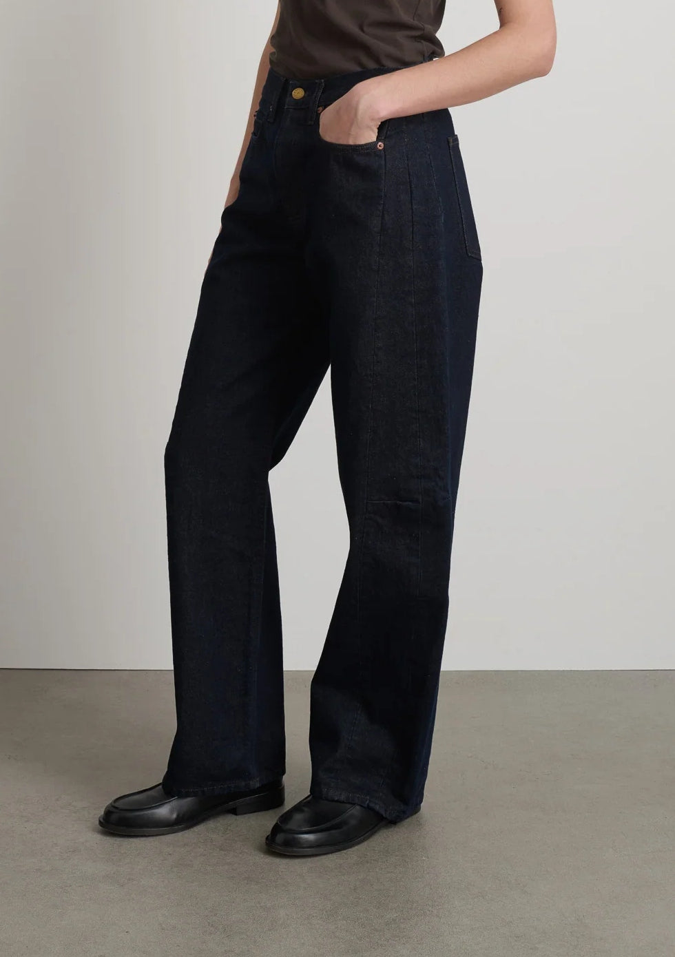 A person wears dark indigo B Sides Slim Lasso Jean in Indigo Brown Rinse wide-leg jeans with curved side seams and black leather loafers.