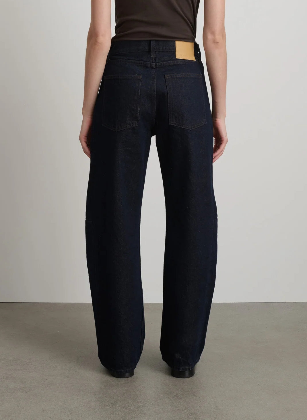 Rear view of the B Sides Slim Lasso Jean in Indigo Brown Rinse, featuring dark blue cotton denim and a tan leather patch on the high-waisted back waistband.