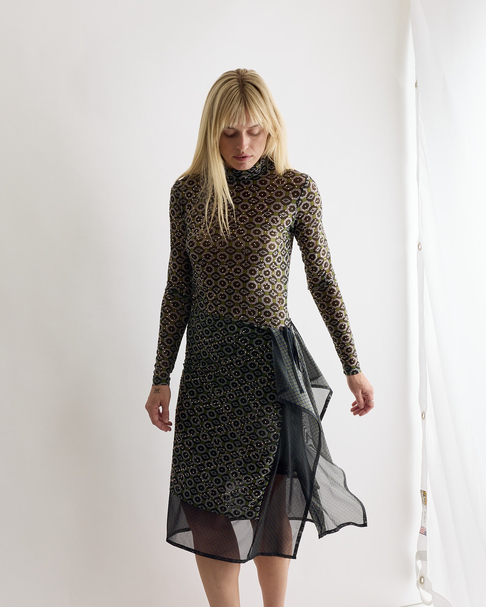 A woman with straight blonde hair wears a Dries Van Noten Embellished Turtleneck Tee in Green, standing against a plain white background with her arms relaxed by her sides and looking down.