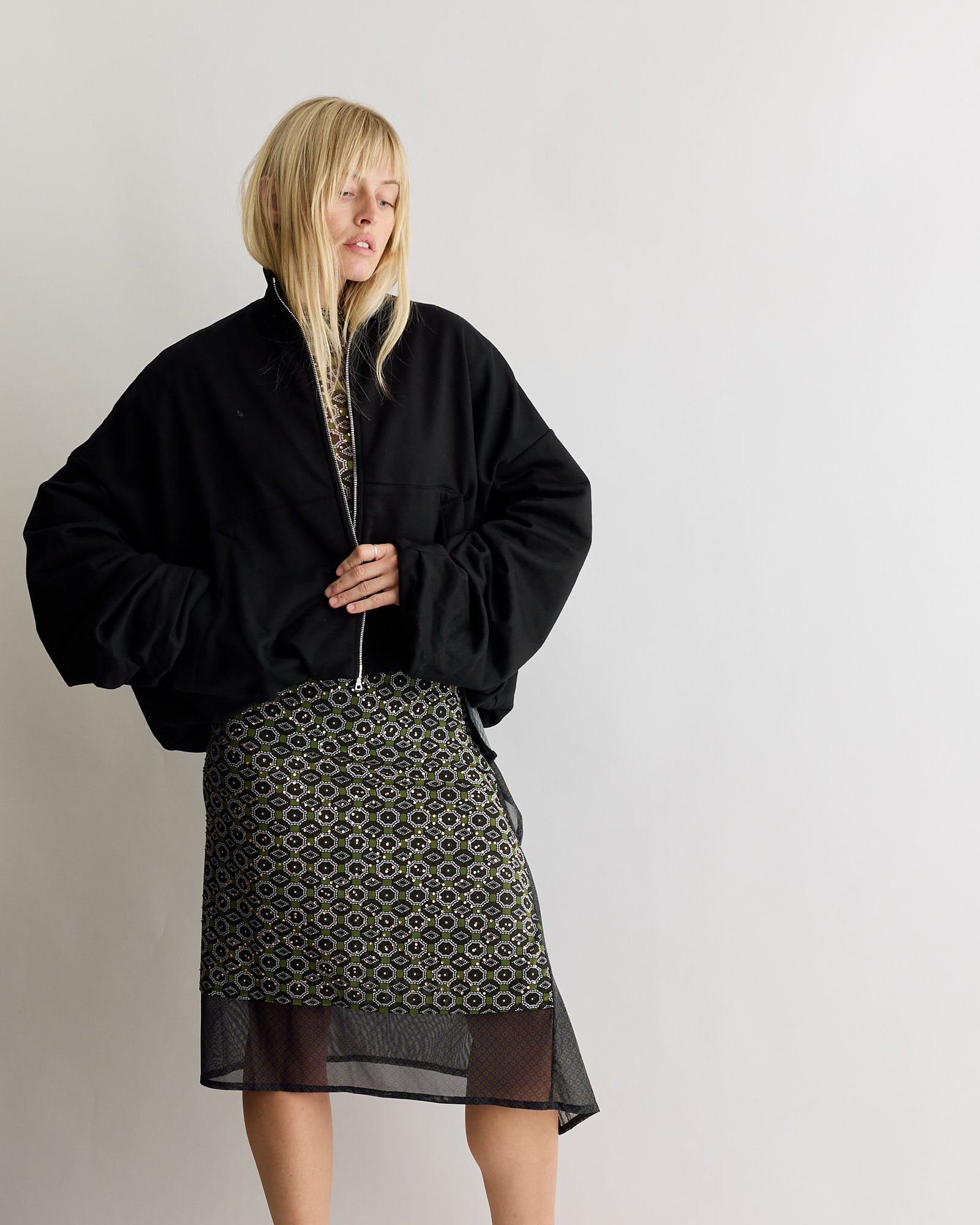 A person with long blonde hair wears the Dries Van Noten Draped Jersey Jacket in Black, styled in an oversized silhouette, and a patterned knee-length skirt, standing against a plain white background.