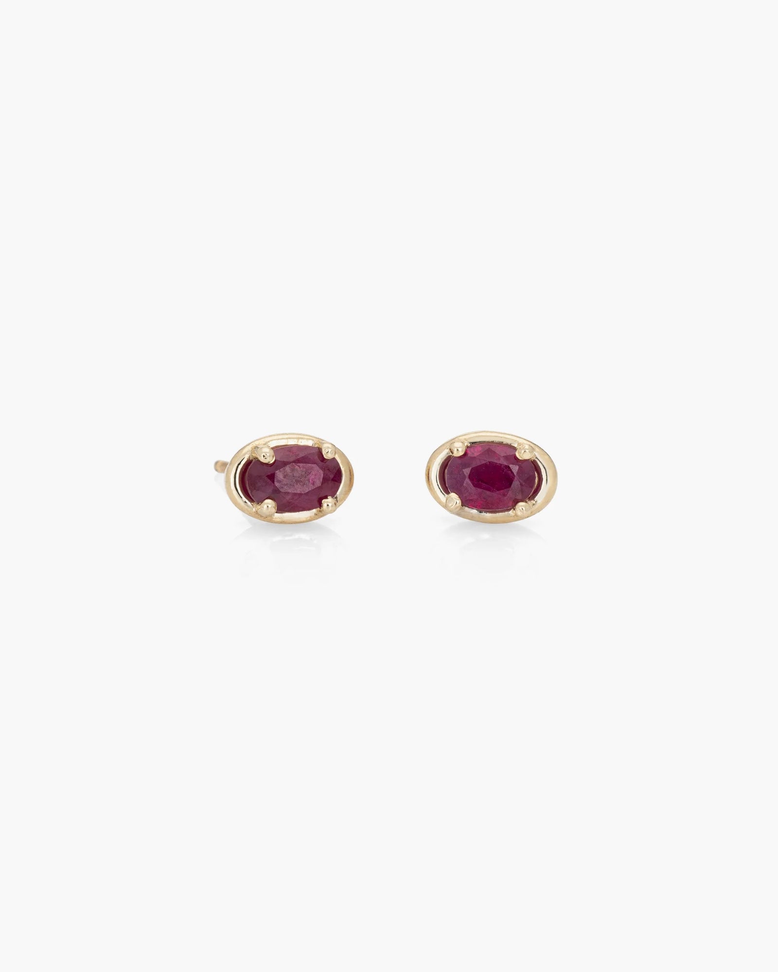 The Kathryn Bentley Ellipse Studs Pair in 14K features small, oval-shaped deep red rubies set in yellow gold prongs, shown against a plain white background.