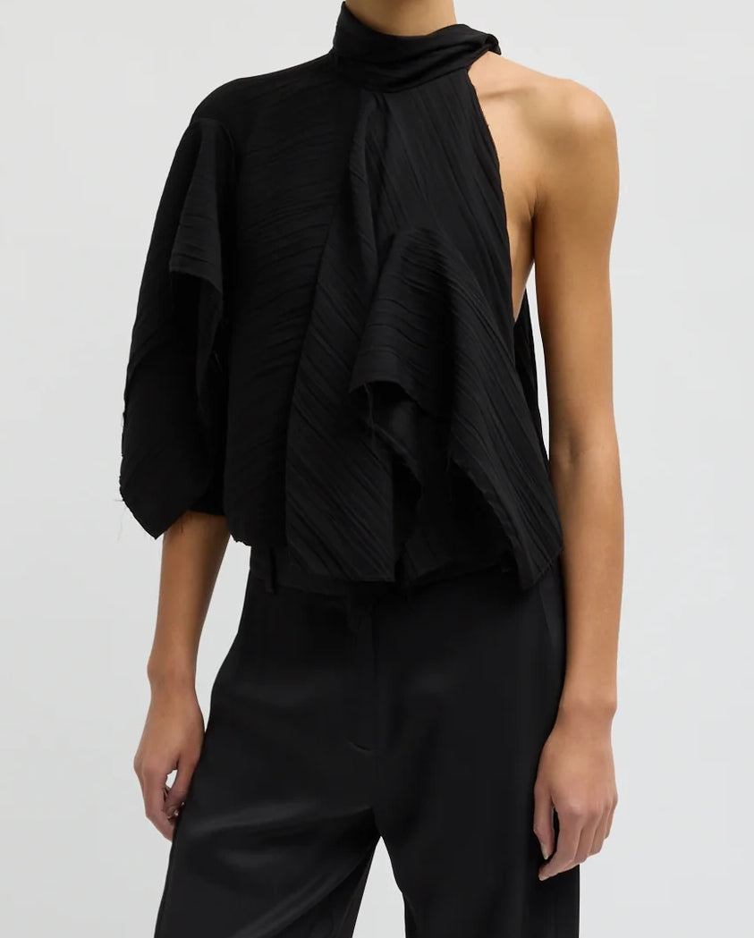 A person wearing Tibi’s Creased Drape Cape Tie Top in Black—a pleated, asymmetrical piece with one sleeveless side and a high neckline—paired with black pants, stands against a plain light background.