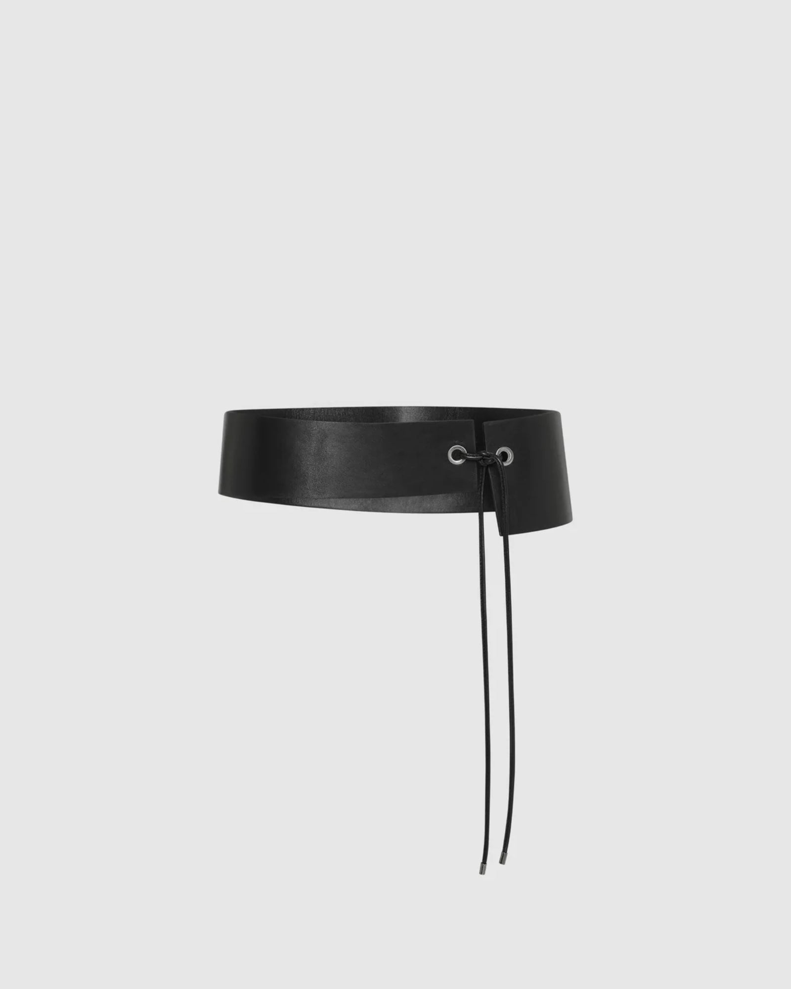 The Asymmetric Belt in Black by ST. AGNI is a wide black leather waist belt with a front lace-up closure featuring black laces, displayed against a plain light gray background.