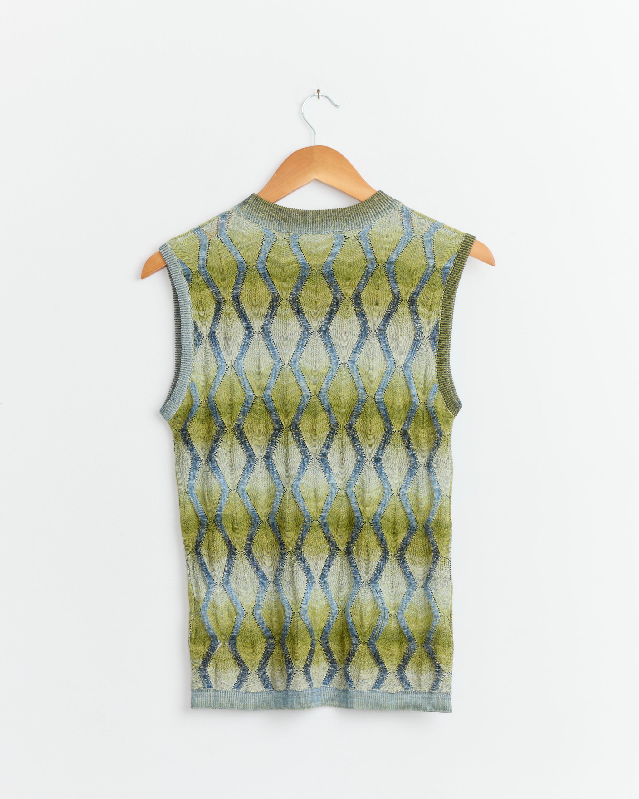 The Henrik Vibskov Resonance Top in Multi Flame, a sleeveless knit vest featuring a green, gray, and blue geometric diamond pattern, hangs on a wooden hanger against a plain white wall.