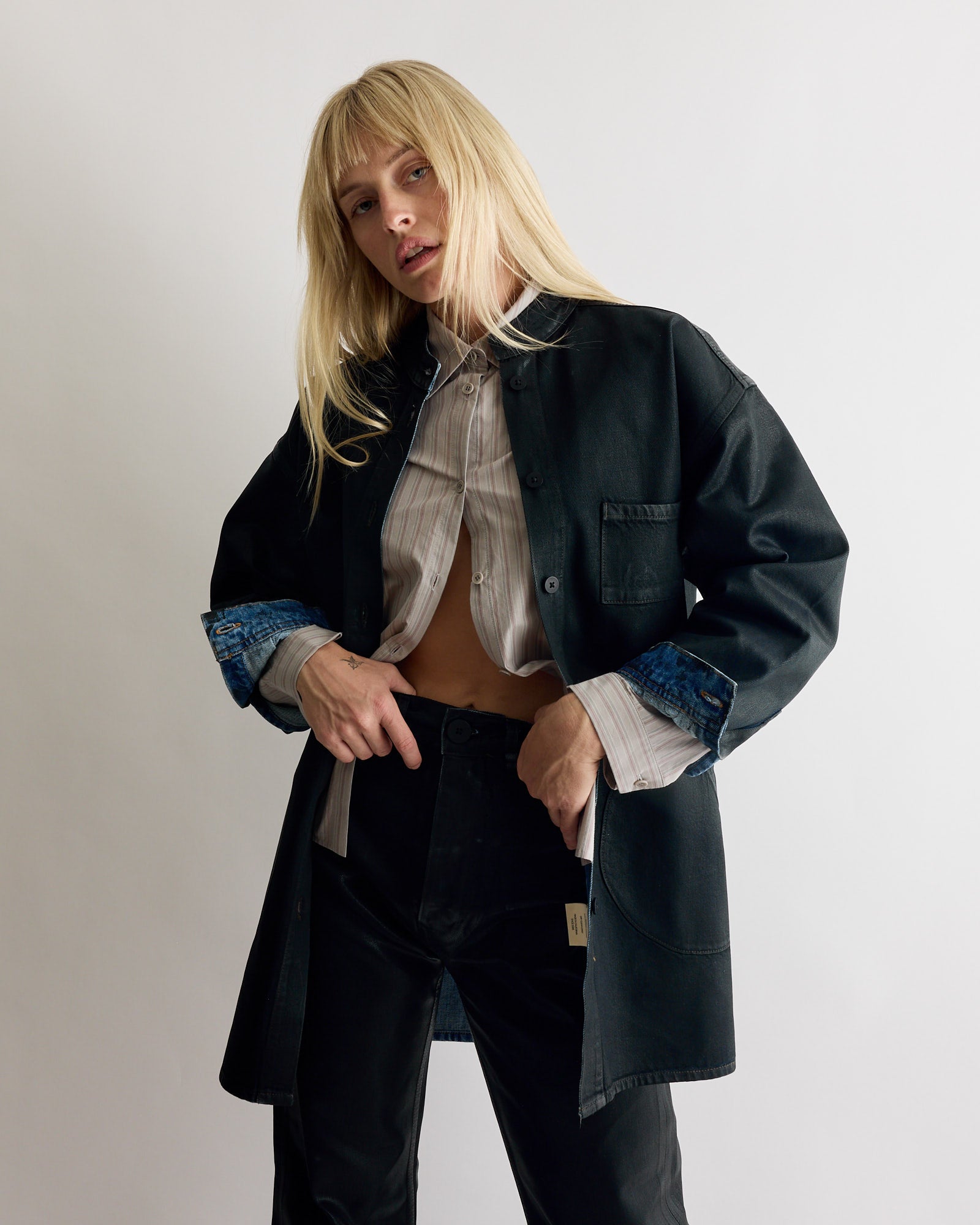 A person with long blonde hair poses against a plain background, wearing Jesse Kamm's Band Collar Okuda Jacket in Black Lacquer over a partially unbuttoned striped shirt and black pants. Their expression is neutral, with hands near their waist.