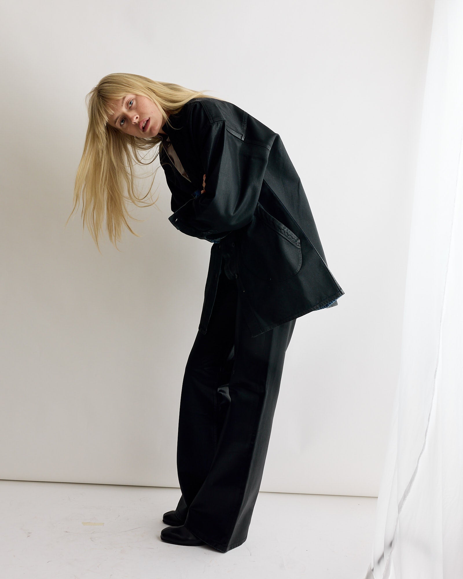A person with long blonde hair wearing an oversized black jacket and the Les Deux Pant in Black Lacquer by Jesse Kamm stands against a plain white background.