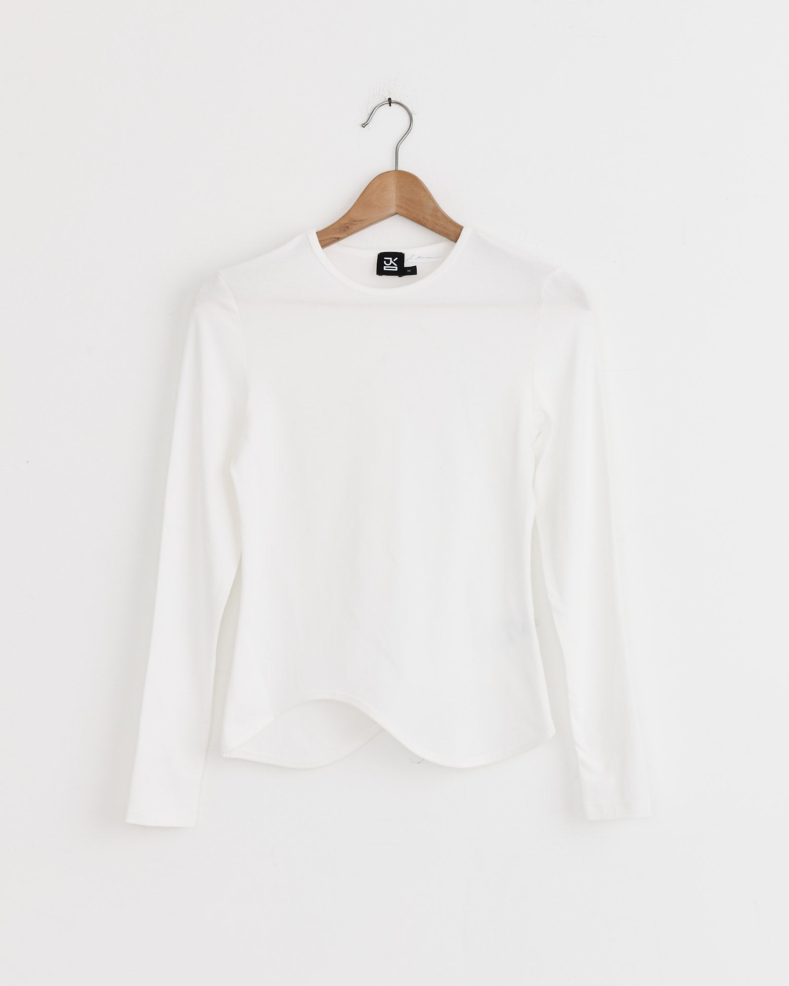 The Ying-Yang Top in White by J. Kim, a minimalist cotton-blend long-sleeve shirt with a round neckline, hangs on a wooden hanger against a white wall.